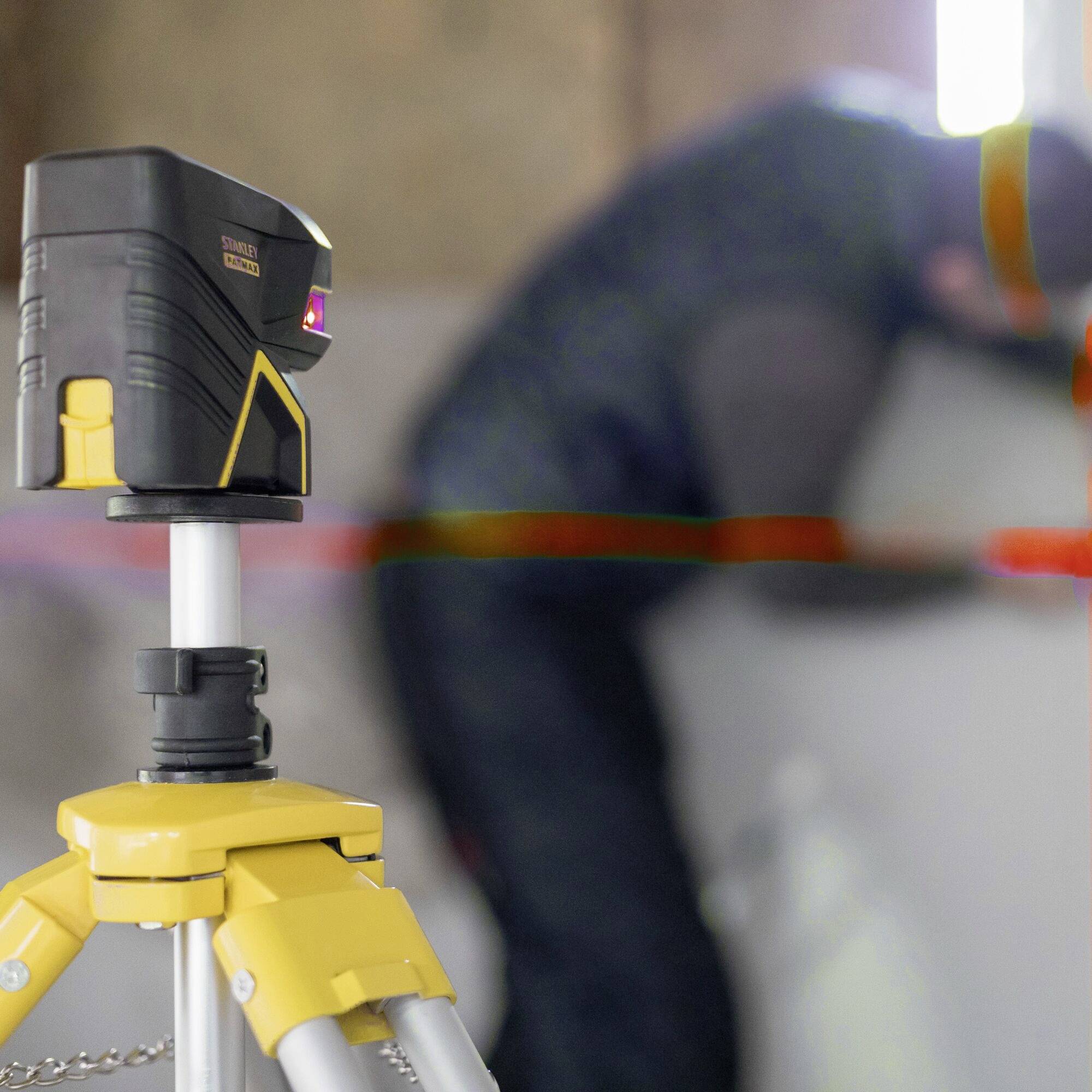 A laser measuring device on a tripod is in focus in the foreground, while a person working on the wall is blurred in the background.