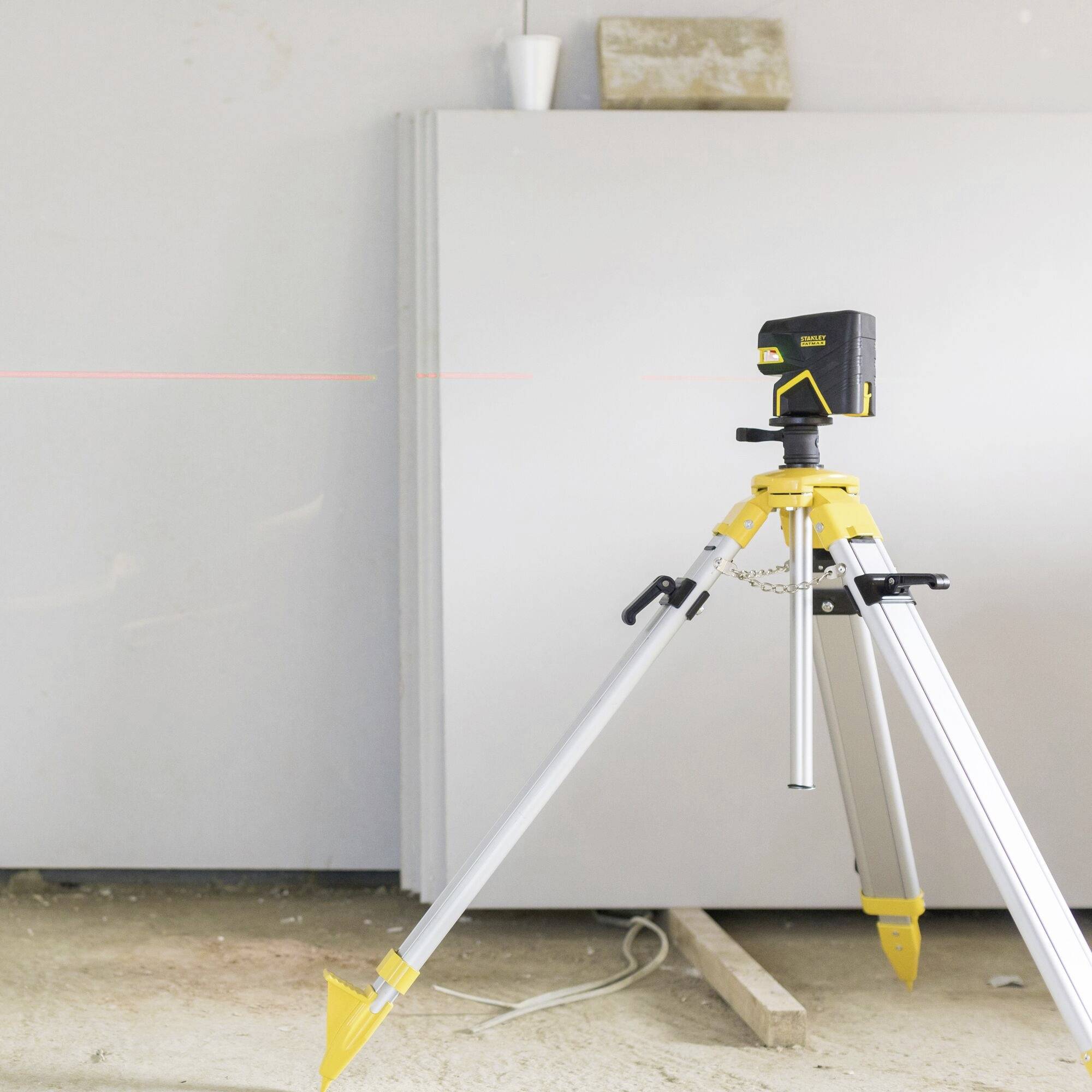 A laser levelling device is mounted on a tripod in a room, projecting a horizontal red line onto the wall for precision measurements.