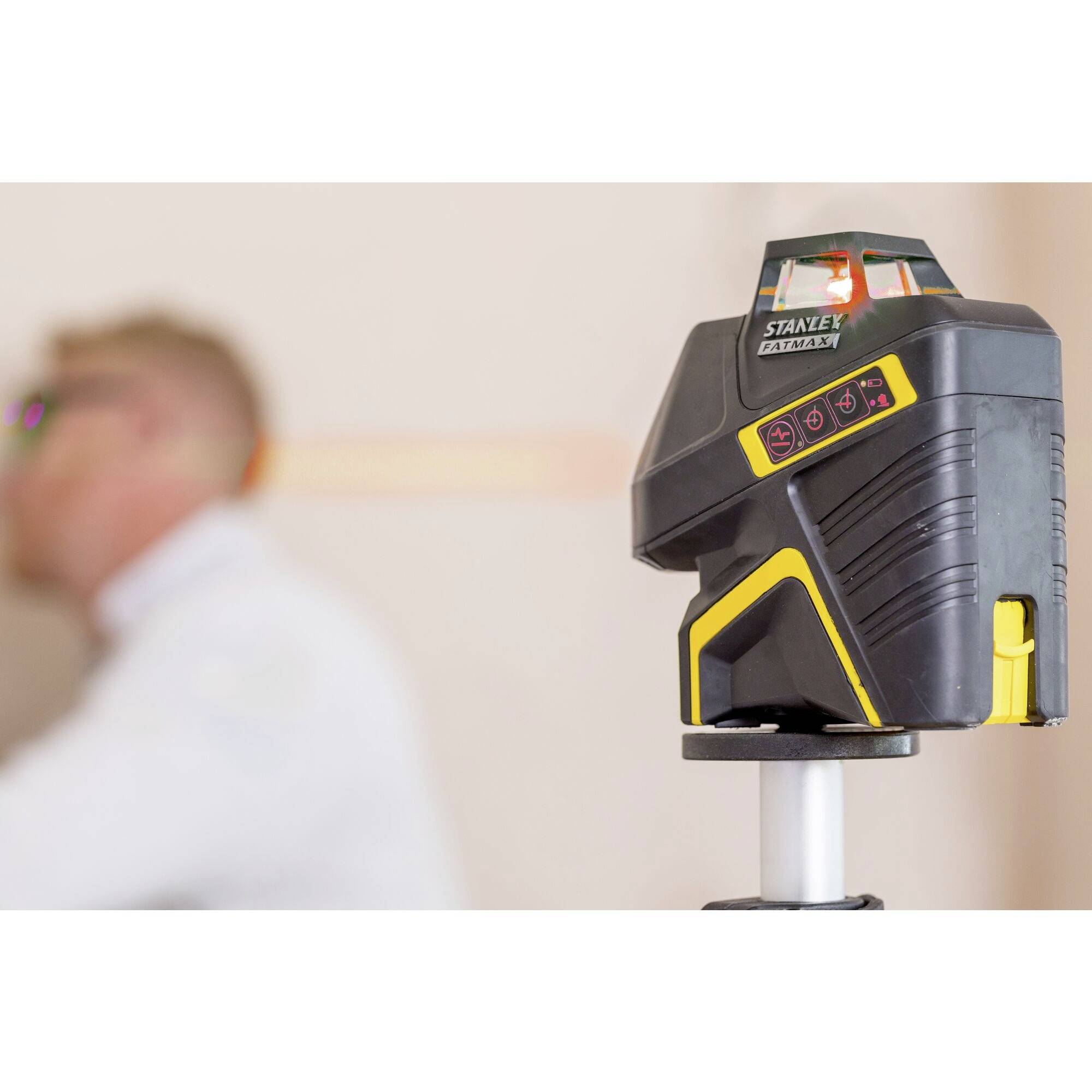 An automatic cross-line laser is mounted on a tripod. In the background, a person wearing white workwear is blurrily visible.