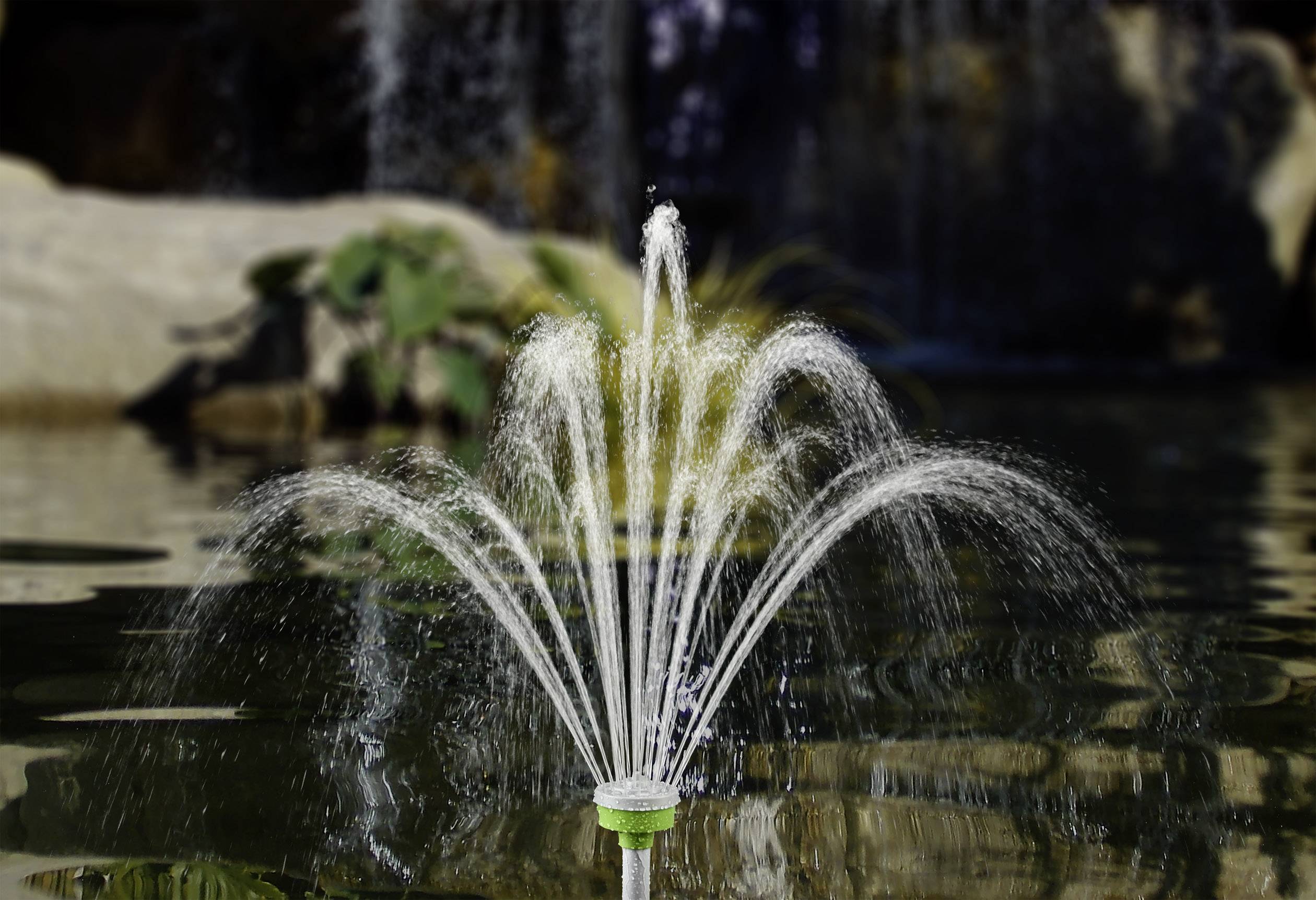 Fountain with arched water jets in a pond; plants and a waterfall visible in the background.
