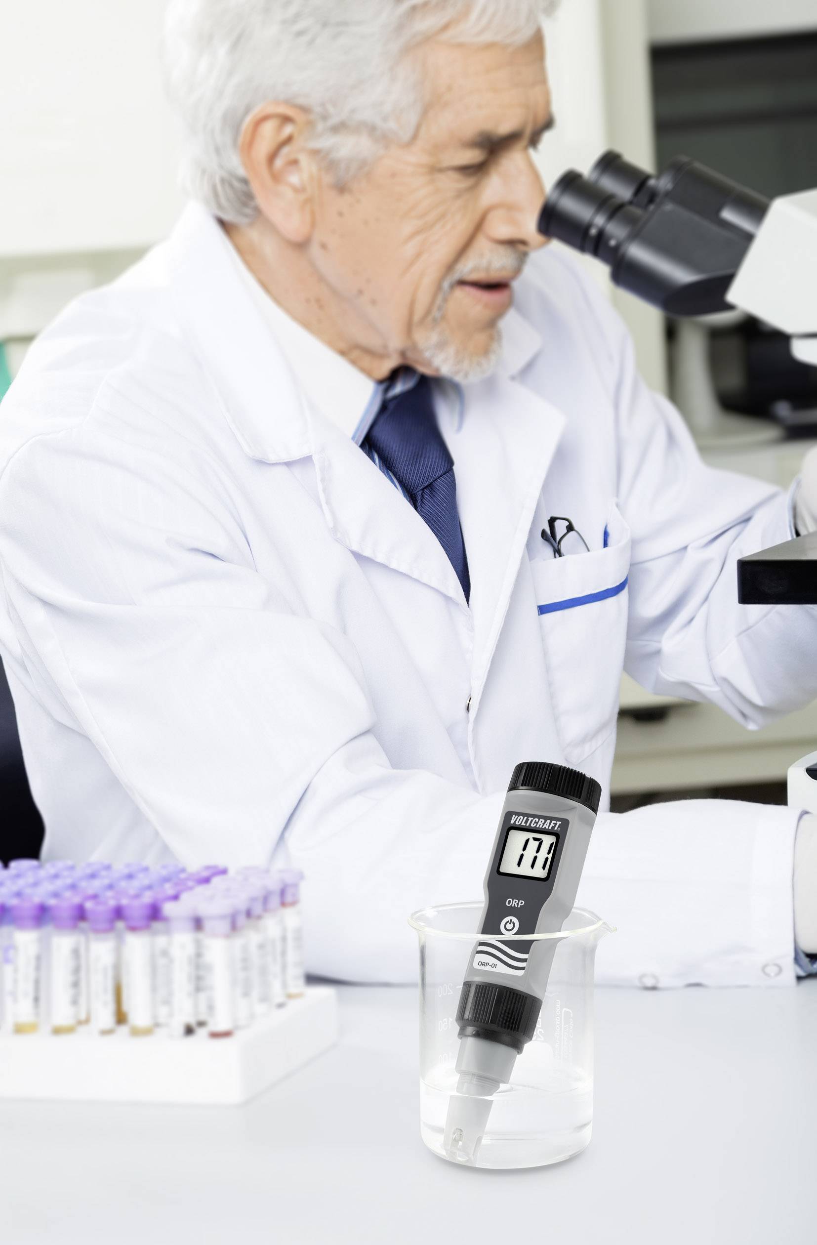 A scientist is examining samples under a microscope in a laboratory. In the foreground, a measuring instrument is placed in a beaker.