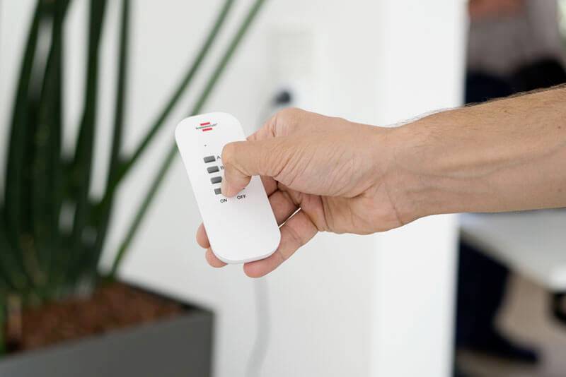 A hand is holding a remote control, pointing at a device. Plant stems and part of a table are visible in the background.