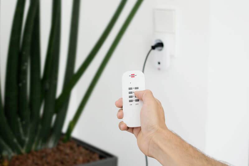 A hand holds a remote control and switches a plug socket on or off. A plant is visible in the background.