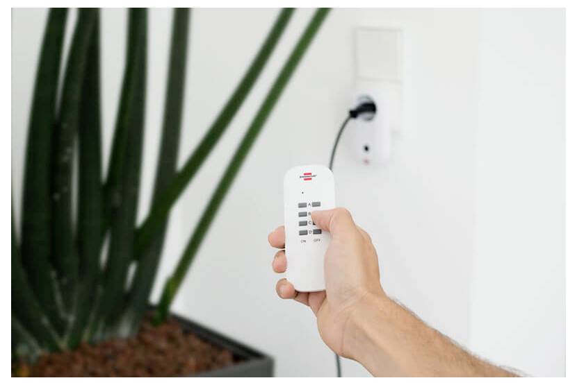 Hand holding a remote control aimed at a wall outlet, turning on a plugged-in device beside a potted plant.<br>