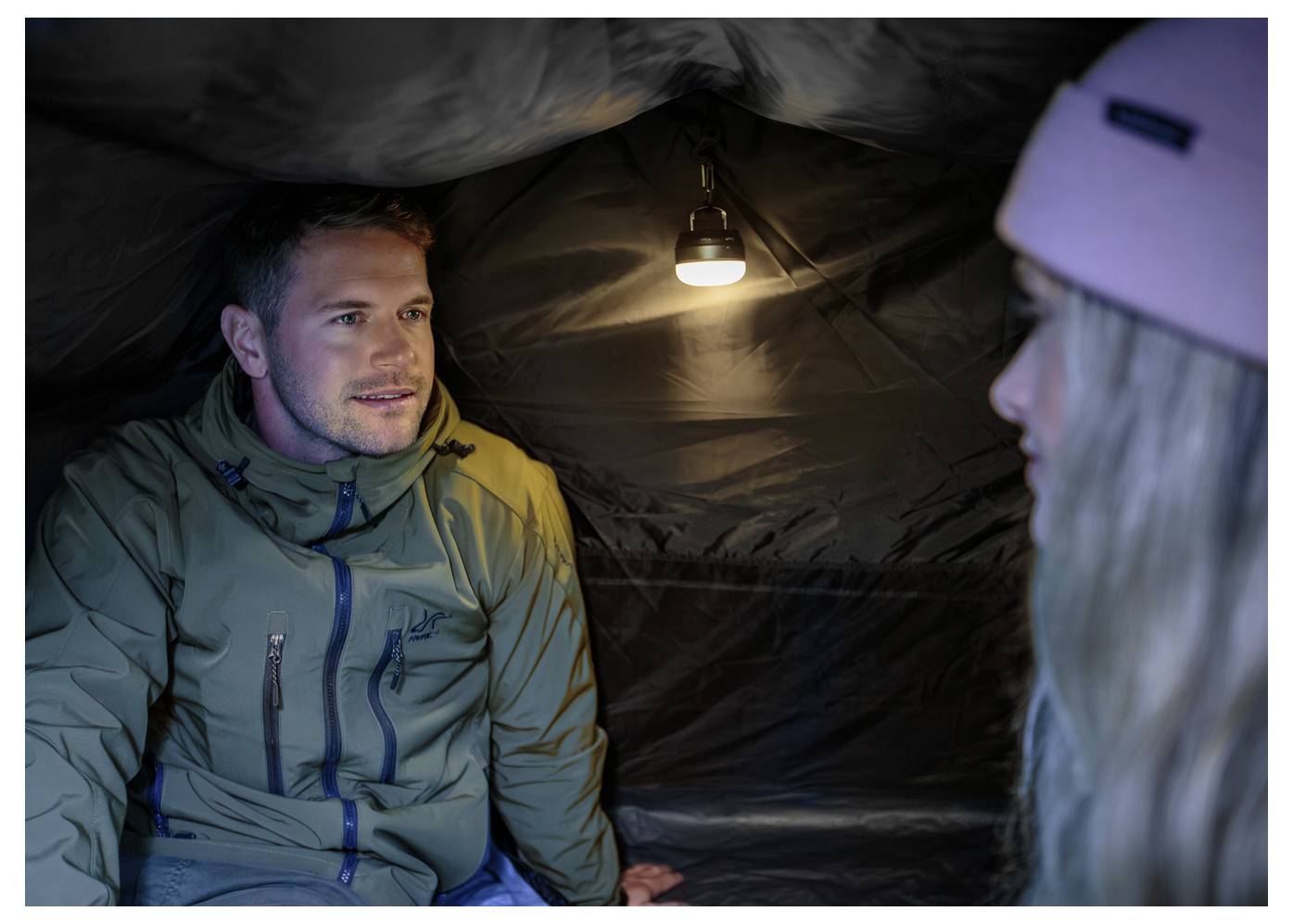 Two people sit inside a small tent lit by a hanging lamp, speaking face to face.