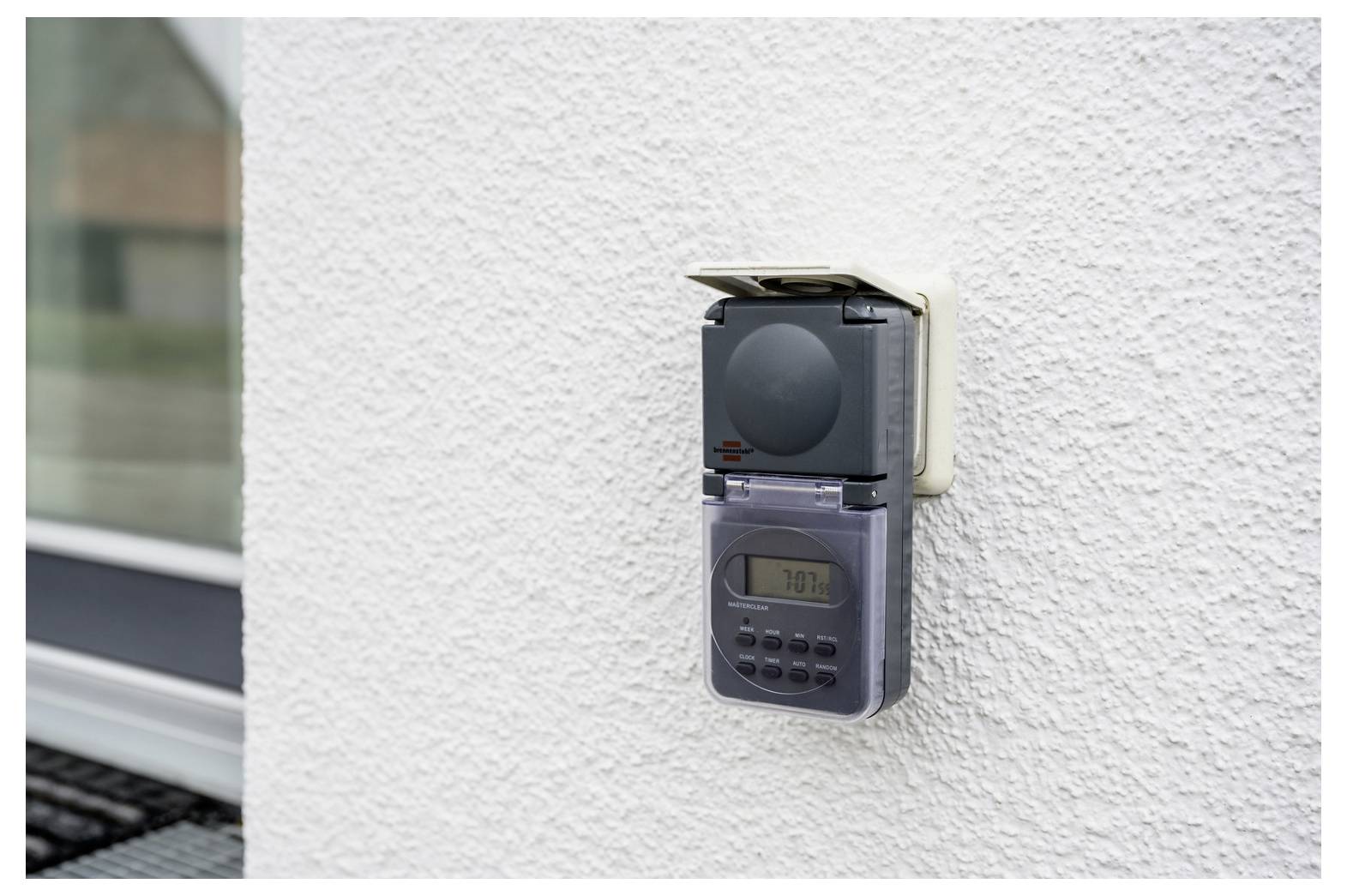 Wall-mounted digital timer or outlet controller on a white textured exterior wall, with a small display and buttons beneath a protective cover.