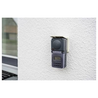 Wall-mounted digital timer or outlet controller on a white textured exterior wall, with a small display and buttons beneath a protective cover.