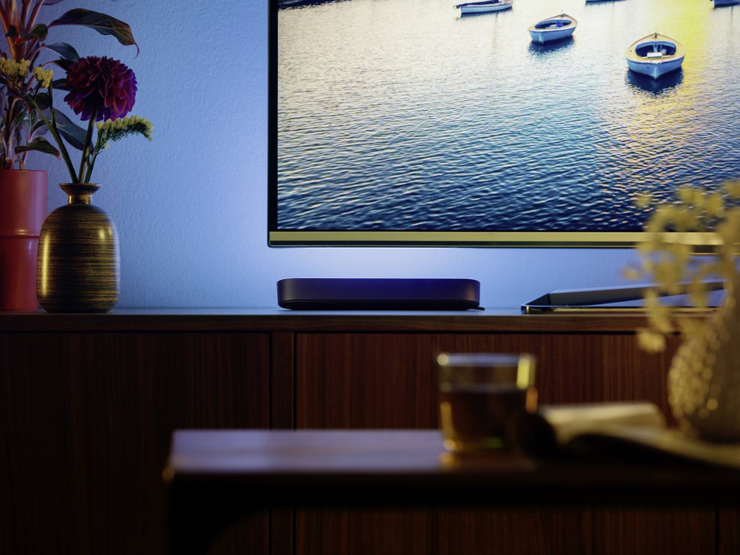 In the foreground, a glass of tea and a book on a table. In the background, a television showing an image of a lake, flowers on a chest of drawers.