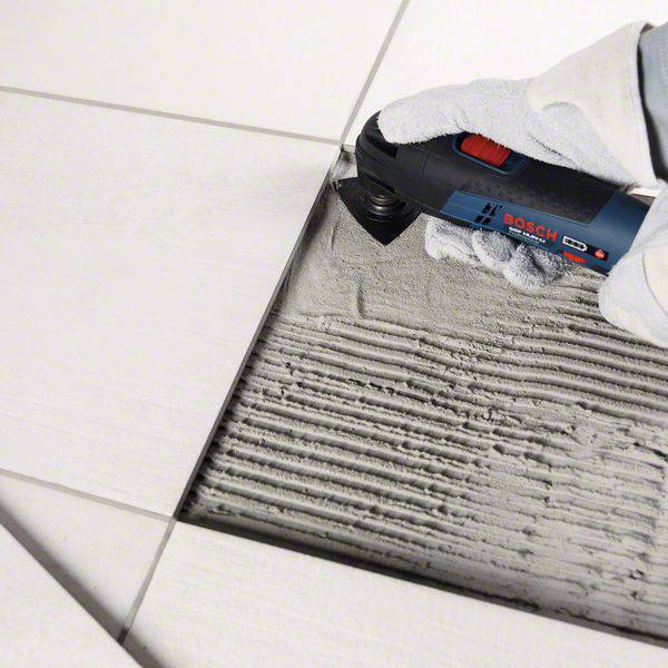A tile is being cut with an electric power tool. The tool is blue and has a safety guard. Tile adhesive is visible.