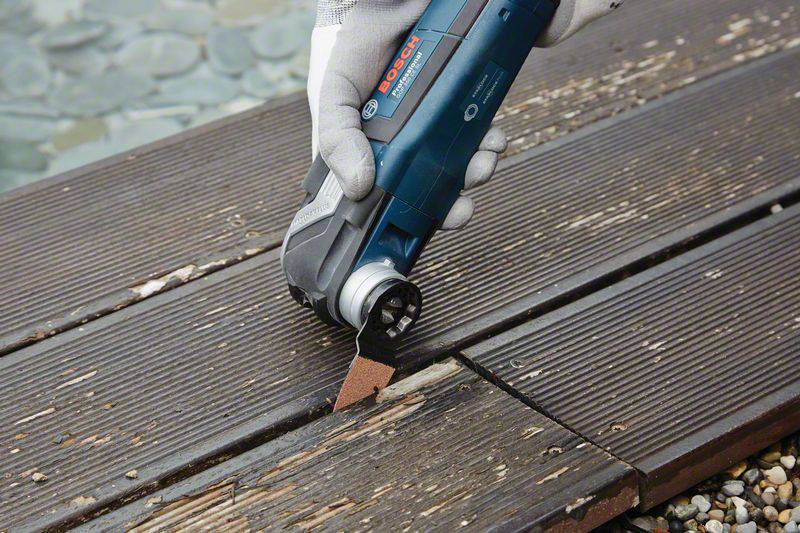 A person is cutting a wooden plank with a blue electric saw from the Bosch brand. The person is wearing white gloves.