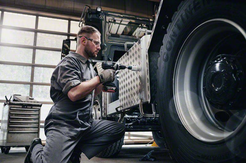A man in workwear is repairing a large vehicle with a cordless drill in a workshop. A tool cabinet is visible in the background.
