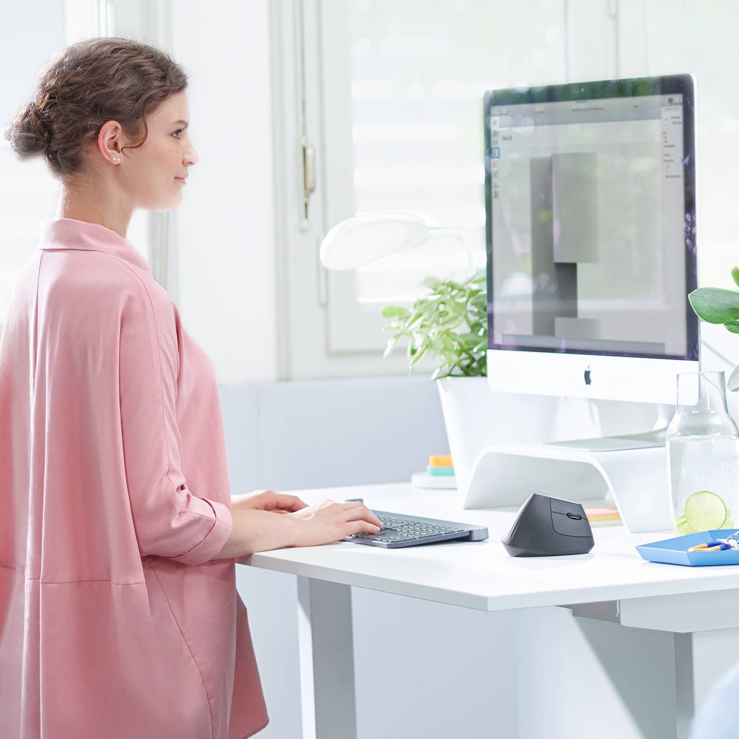 A woman is working on a computer in a modern office. Plants and drinks are on the table, and light is streaming through the window.