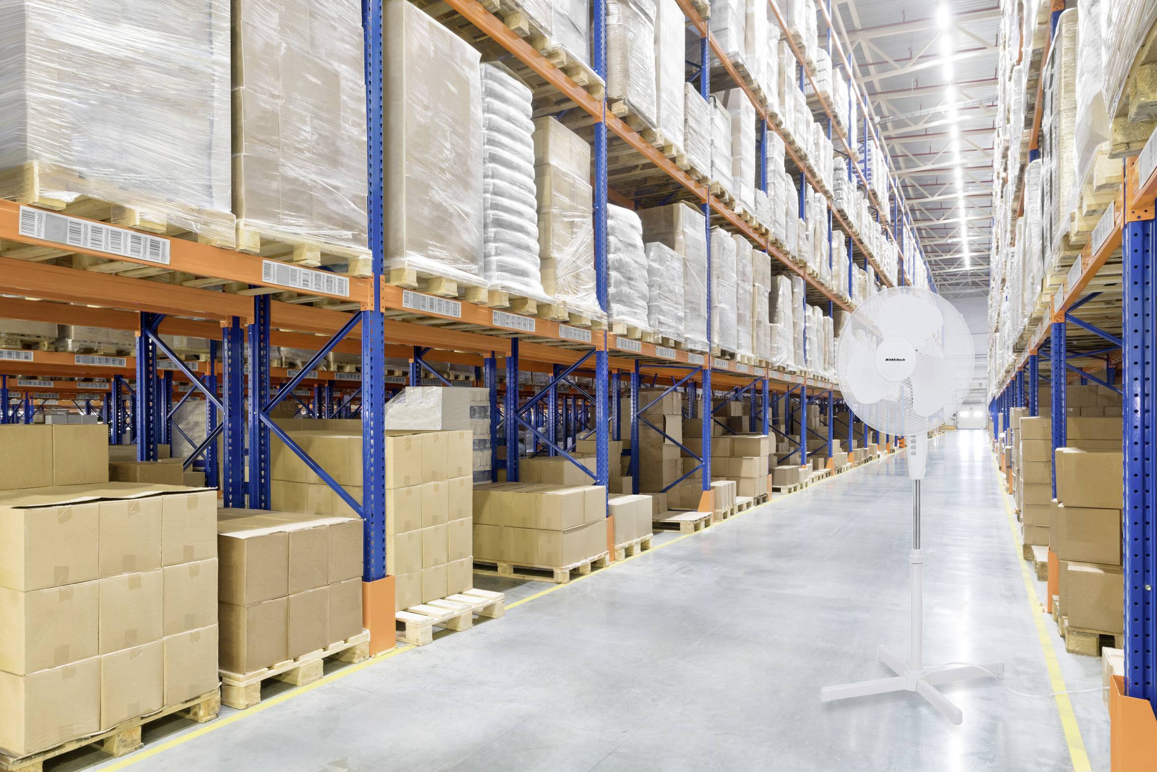 A large warehouse with stacked boxes on blue shelving. The floor is made of concrete, with a fan positioned on the right side of the image.