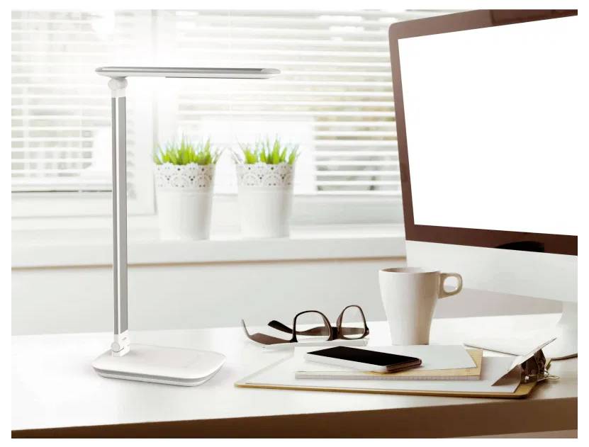 'A modern desk setup with a sleek lamp, a computer monitor, a white coffee cup, eyeglasses, and a notepad on a light-colored table.'