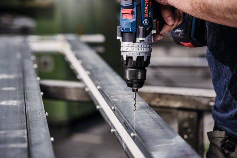 A hand holds an electric drill and is drilling into a metal profile. The drill is blue and features the logo of a tool brand.