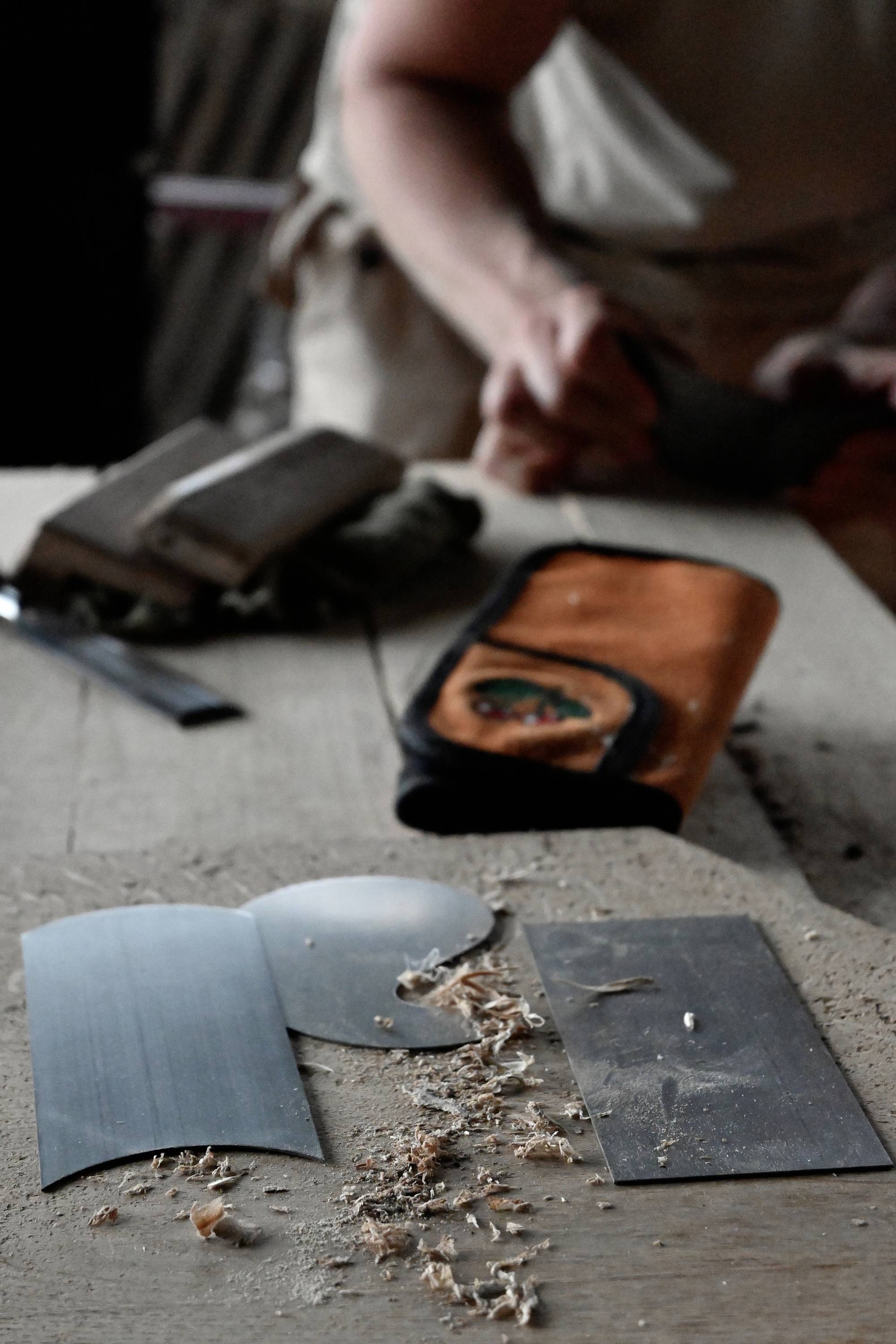 A person is working in a woodworking workshop. Wood shavings, a sharpening tool, and an orange bag are lying on a table.