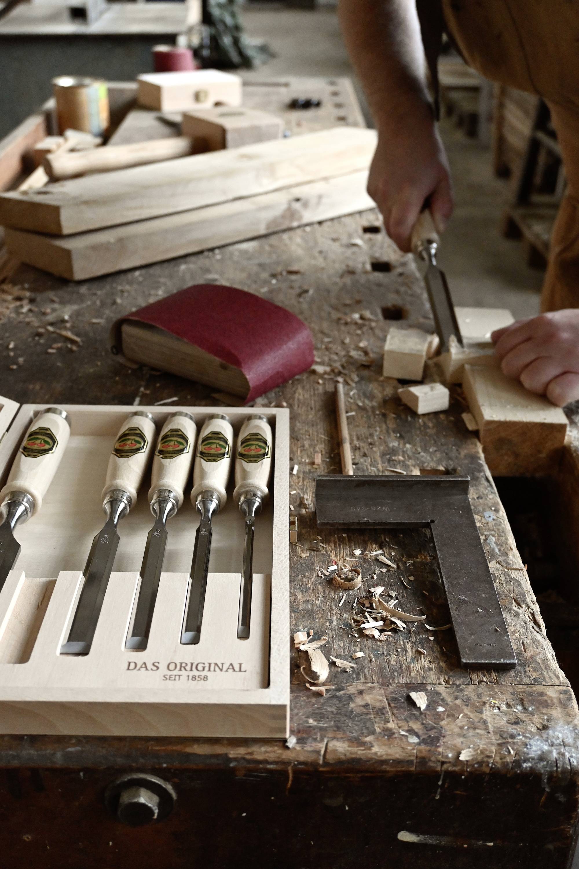 A carpenter is working wood with a chisel on a workbench. Beside him are additional chisels in a box and pieces of wood.