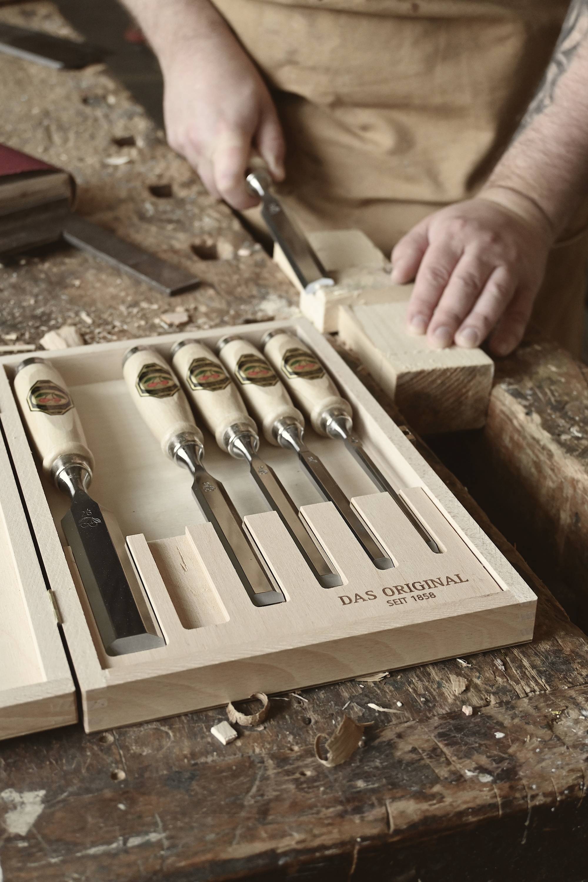 A craftsman is carving with a wood chisel from a set of five chisels, which are carefully laid out on a work surface.