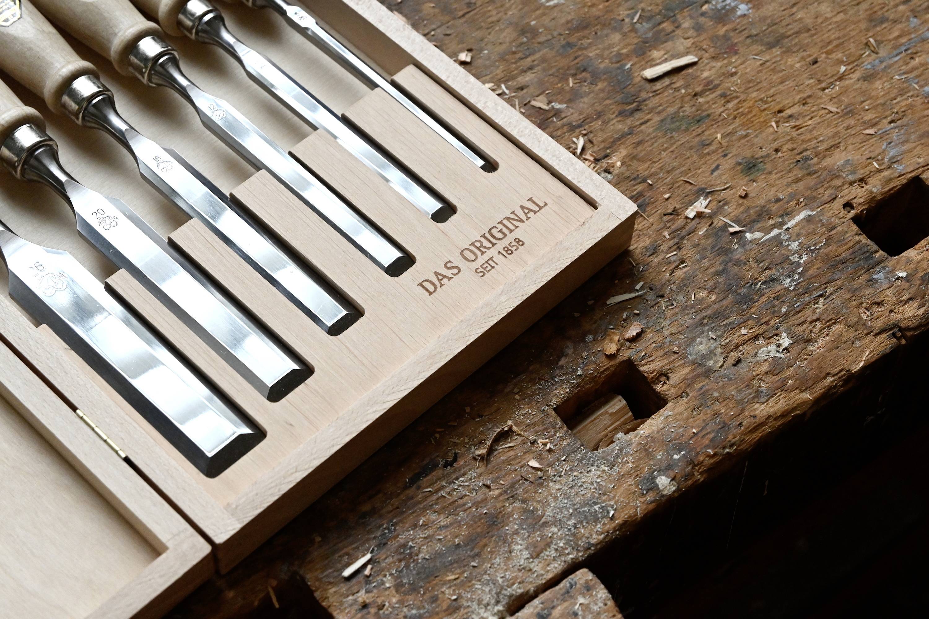 Wooden workbench with woodcarving tools in an open box. On the box it says 'THE ORIGINAL SINCE 1858'. Tools are carefully arranged.