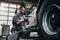 A mechanic in workwear is repairing a large vehicle with an electric tool in a workshop.
