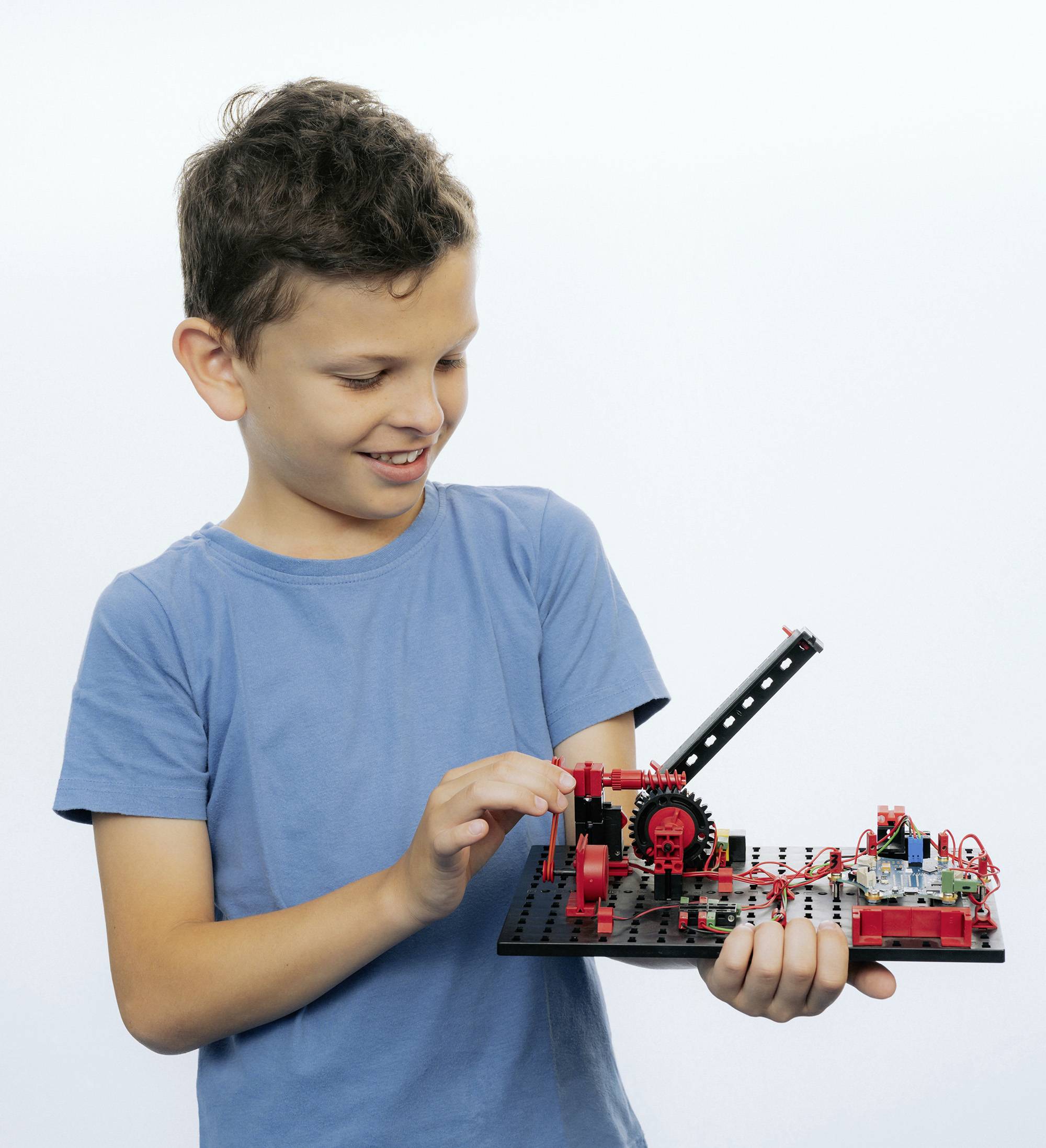 A boy in a blue T-shirt is holding and examining a technical model he has built using red and black components.