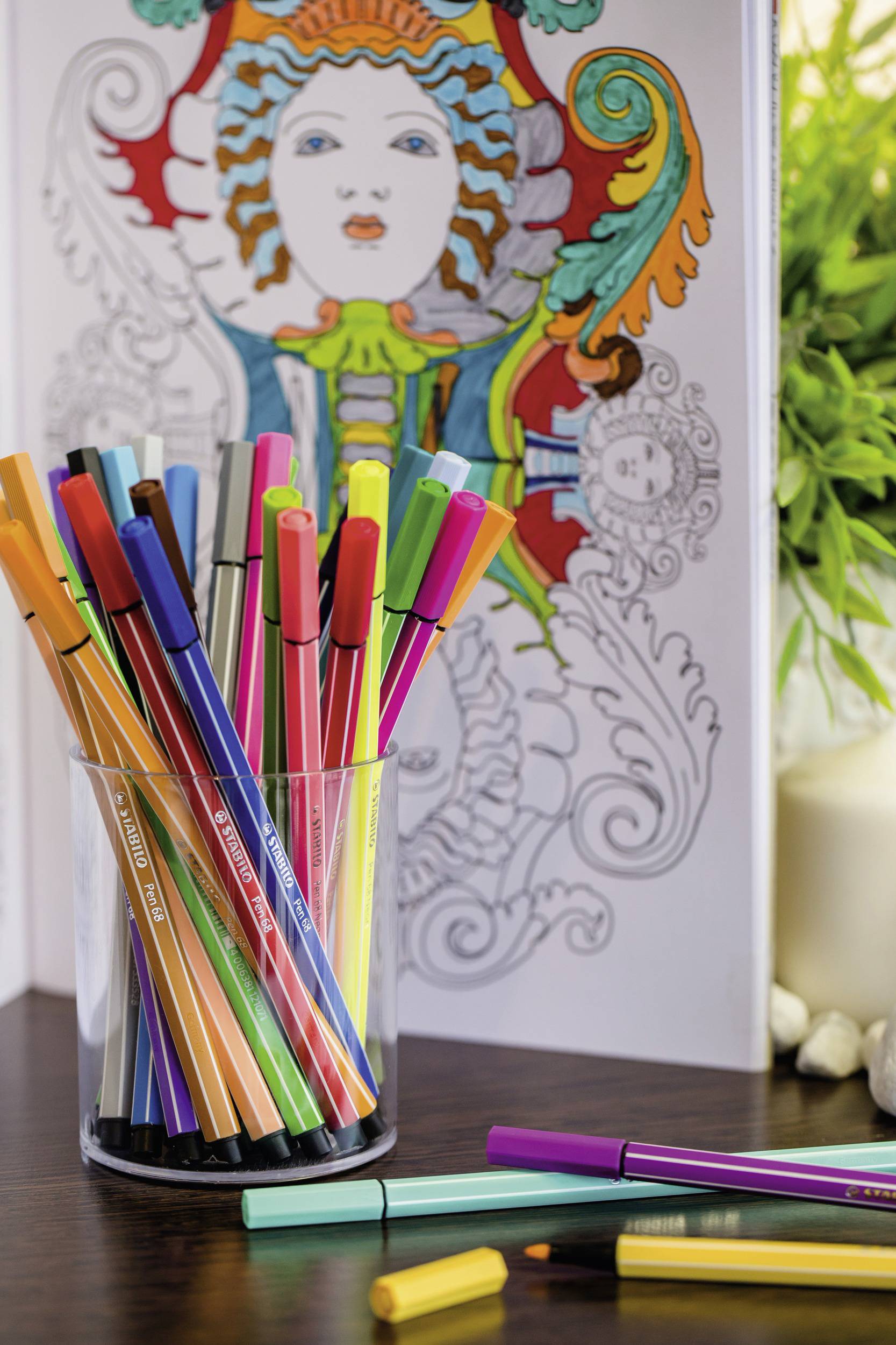 Colourful felt-tip pens stand in a glass in front of a partially coloured colouring book with an artistic, vibrant design.