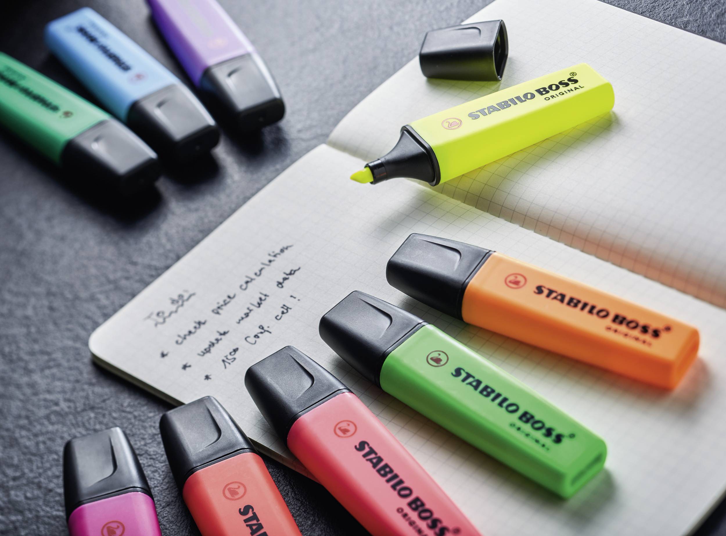 Multicoloured highlighter pens from the Stabilo brand are lying next to a notebook. One highlighter has its cap removed and is marking text.
