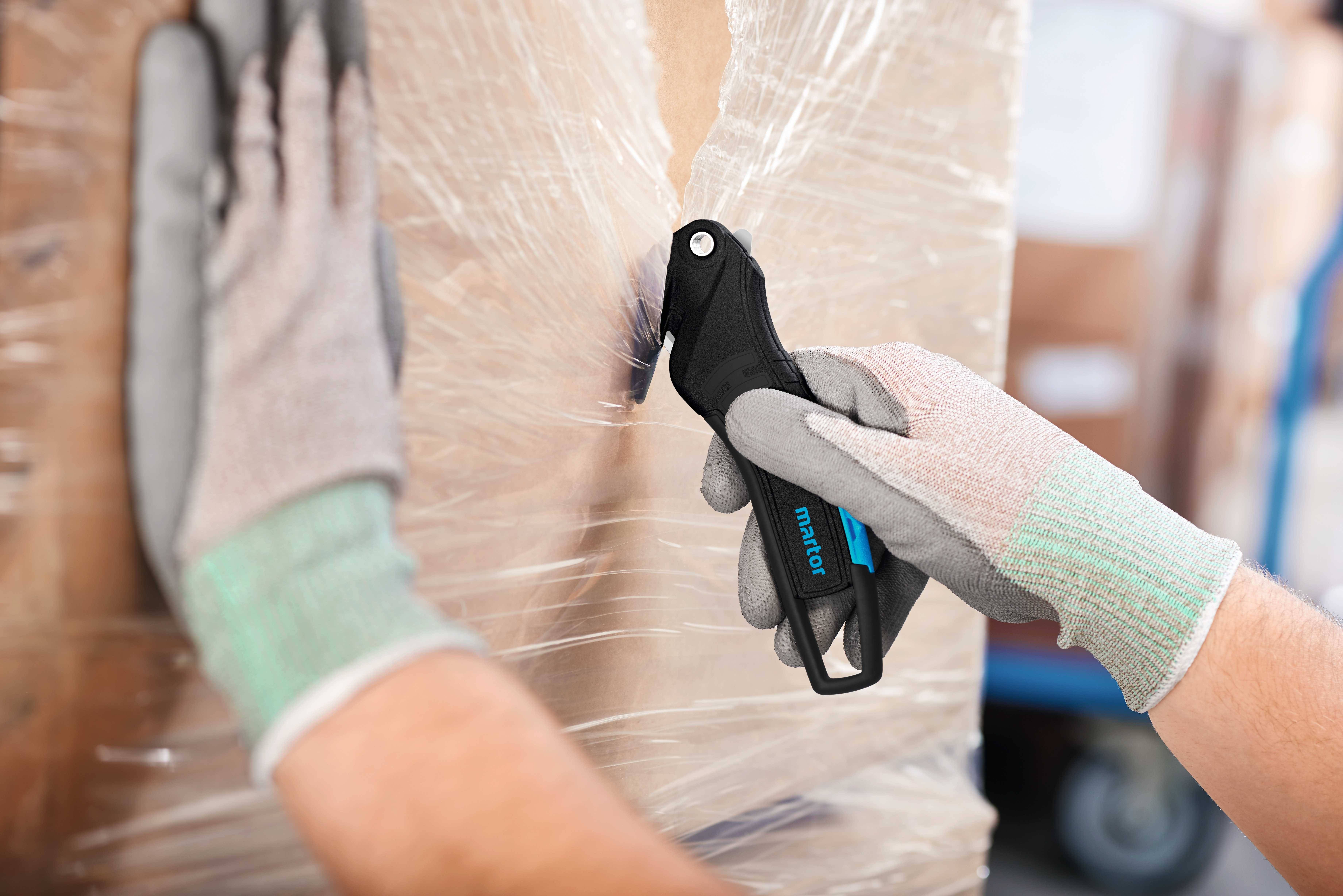 A person is cutting packaging film from a cardboard box using a safety knife. The person is wearing work gloves.