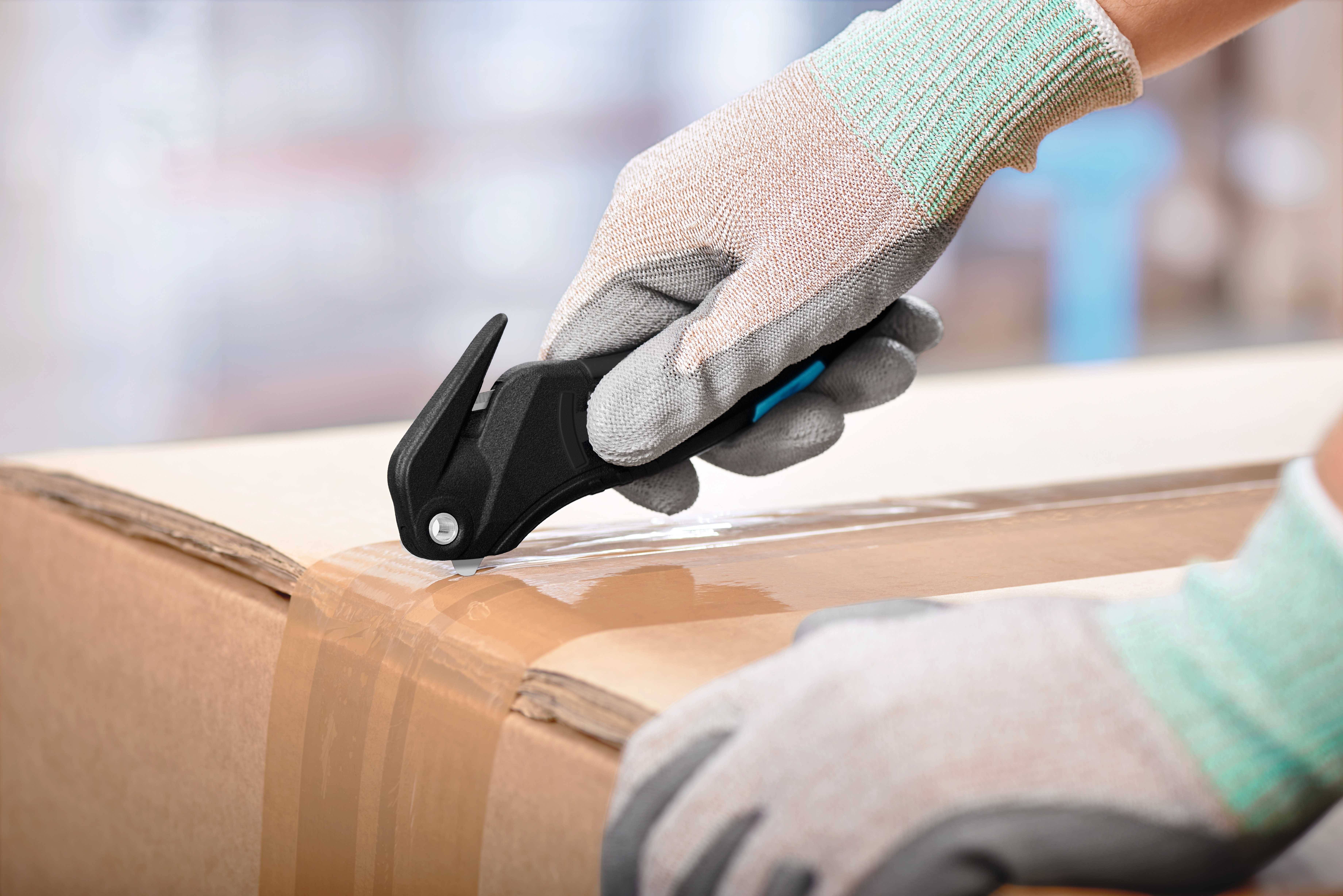 A person wearing work gloves is using a safety knife to cut tape on a cardboard box.