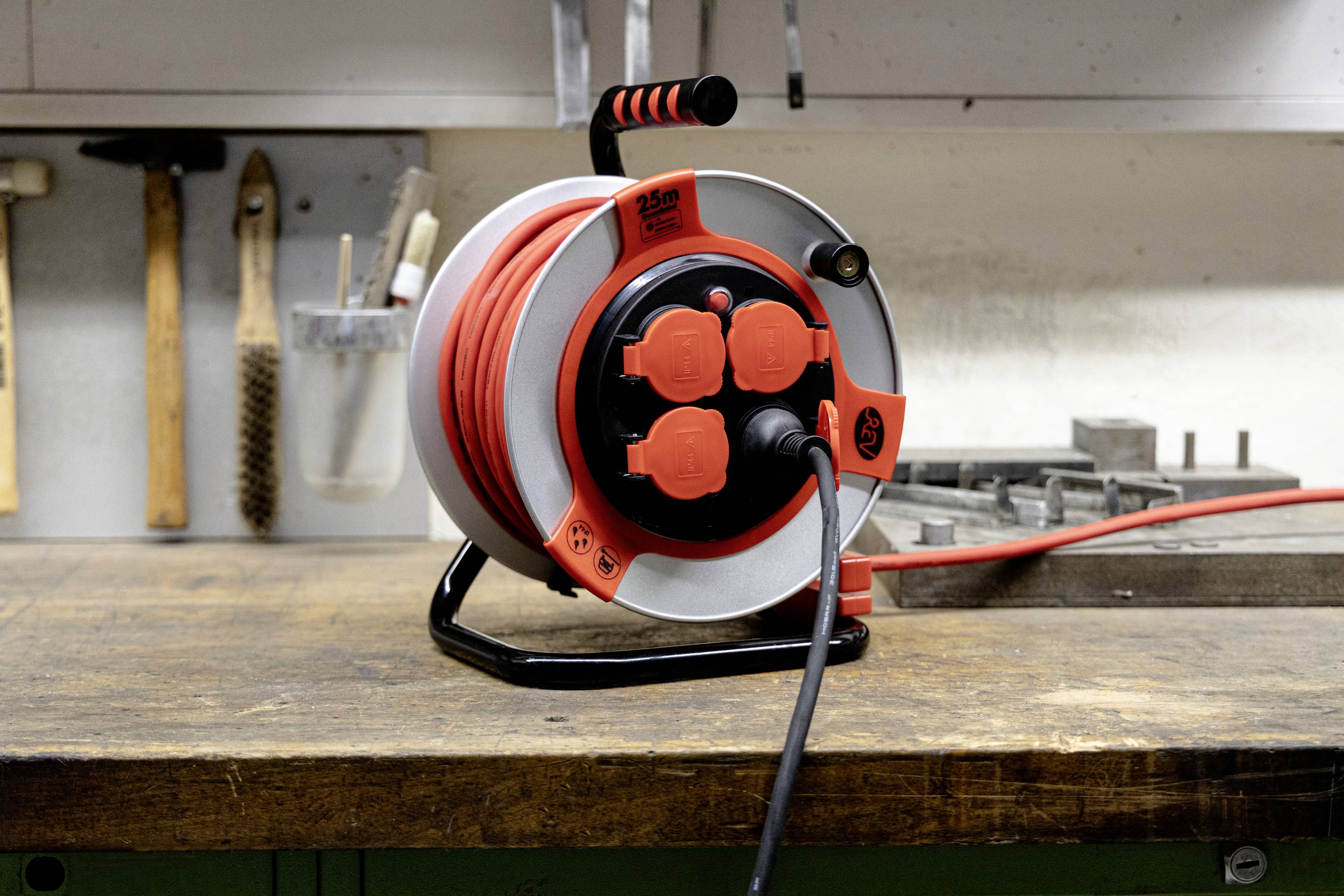 A red cable reel with multiple plug sockets sits on a workbench in a workshop. The background shows tools and screws.