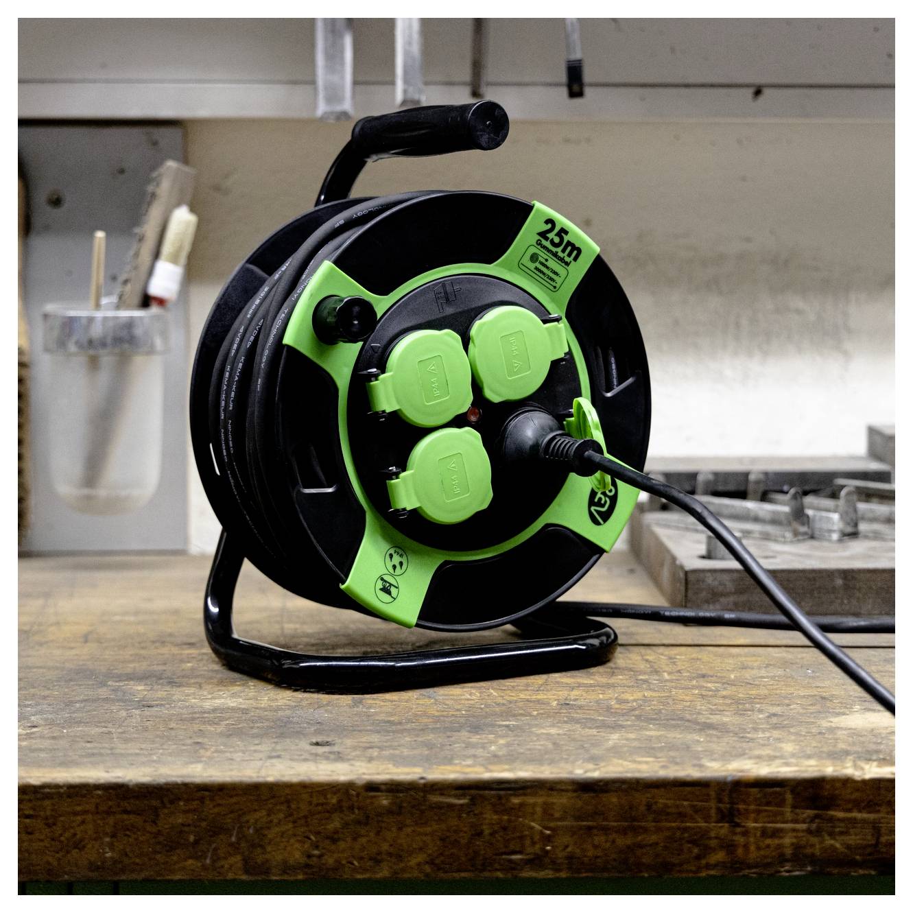 A green and black extension cord reel sits on a wooden workbench. The reel is marked '25m' and has multiple outlets. A cord is plugged in.
