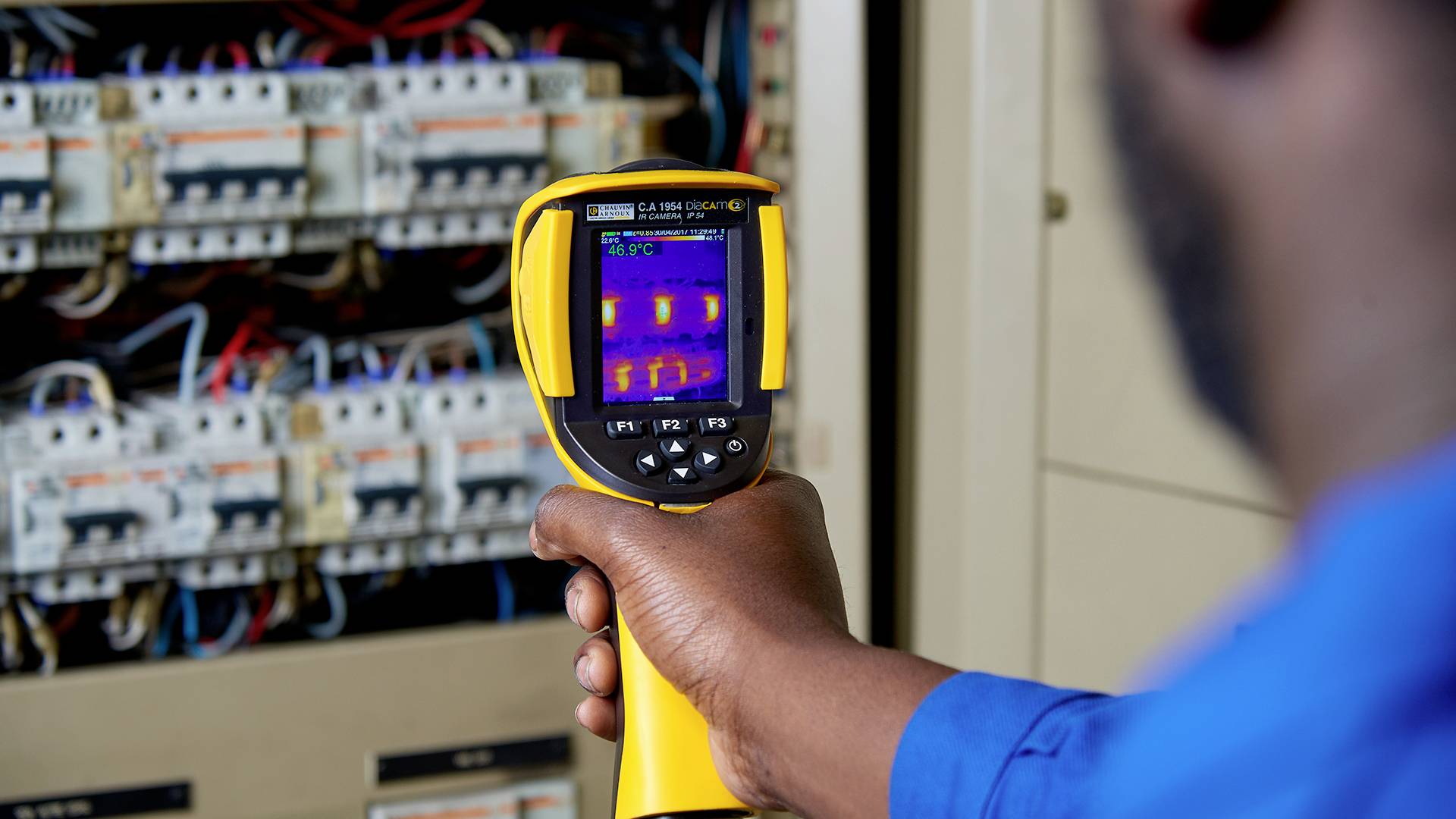 A person is using a thermal imaging camera to check electrical junction boxes for hot spots. The screen displays several coloured points.