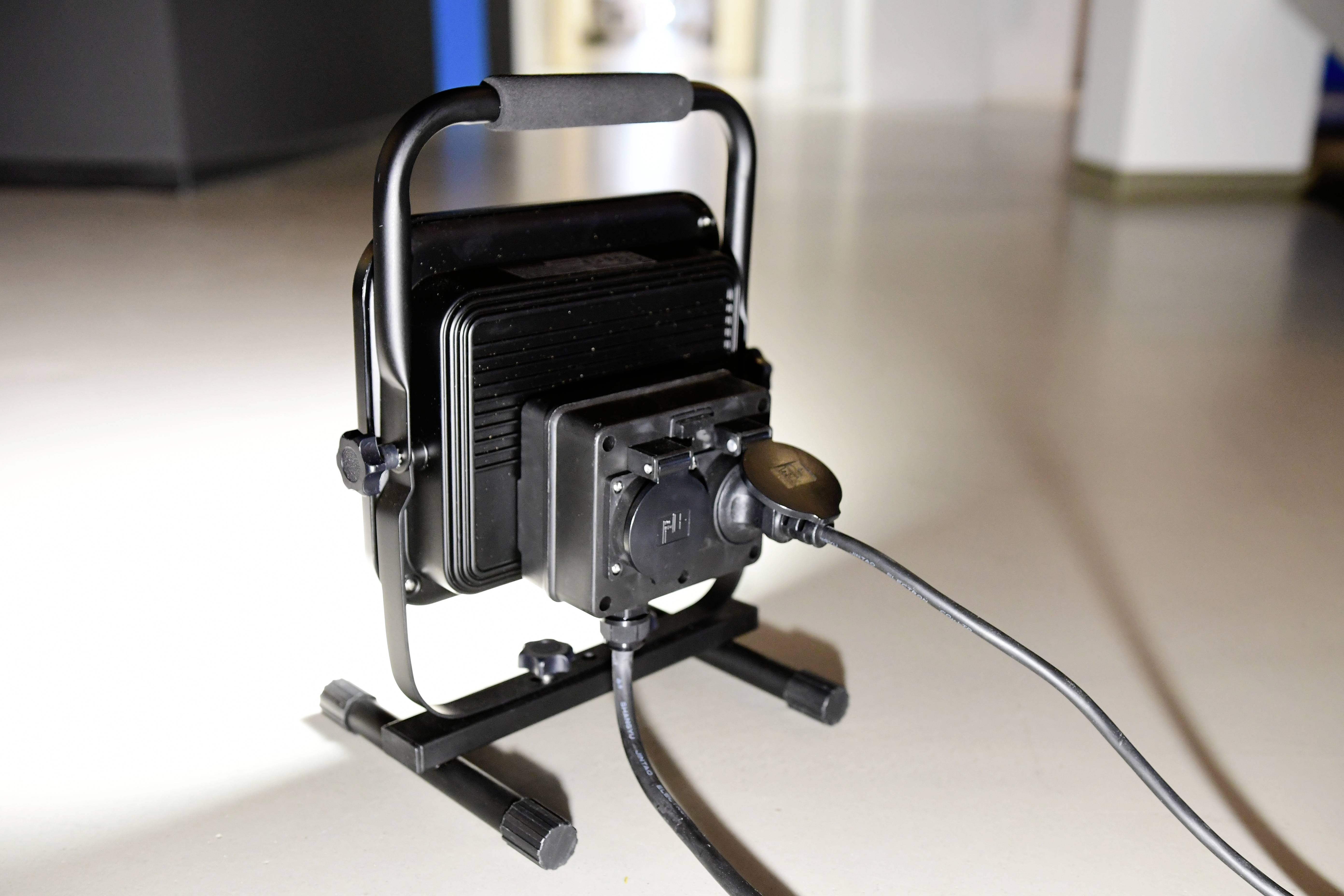 A black work light with a power cable is standing on a floor in a room. The light is switched on but not illuminating.