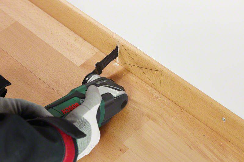 A hand wearing a glove uses an electric multi-tool to cut a piece of decorative trim on the floor.