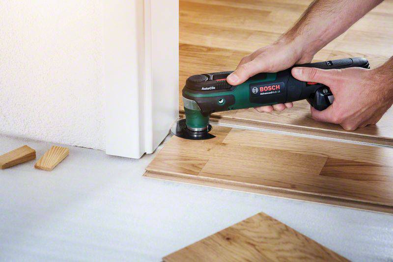 A person is cutting laminate flooring with an electric multi-tool at a doorway. Wooden pieces are lying on the floor.
