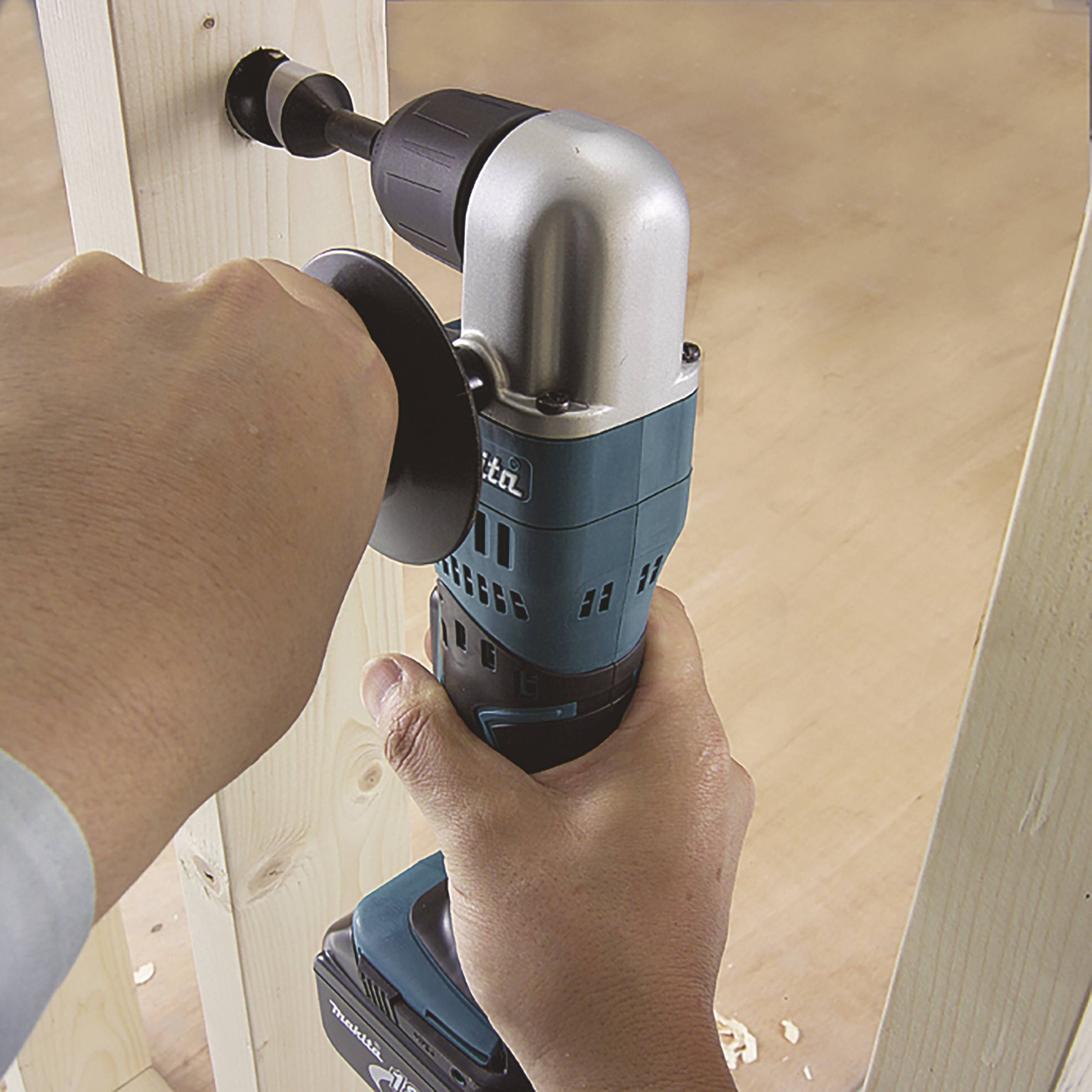 A person is drilling a large hole into a wooden post using a blue cordless drill. Wood shavings are scattered on the ground.