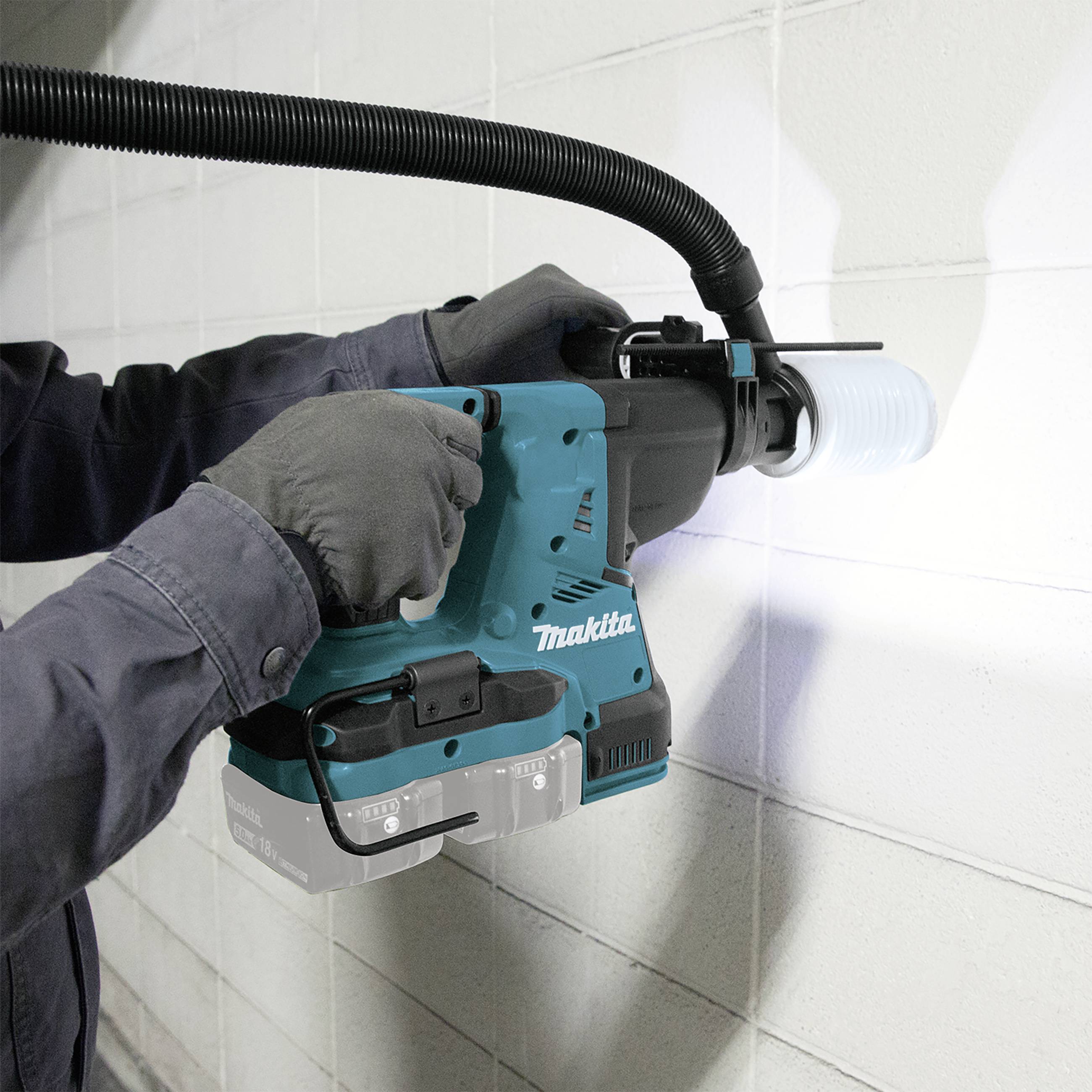 A person is using a blue hammer drill on a white wall. A vacuum cleaner adapter removes dust during the work.