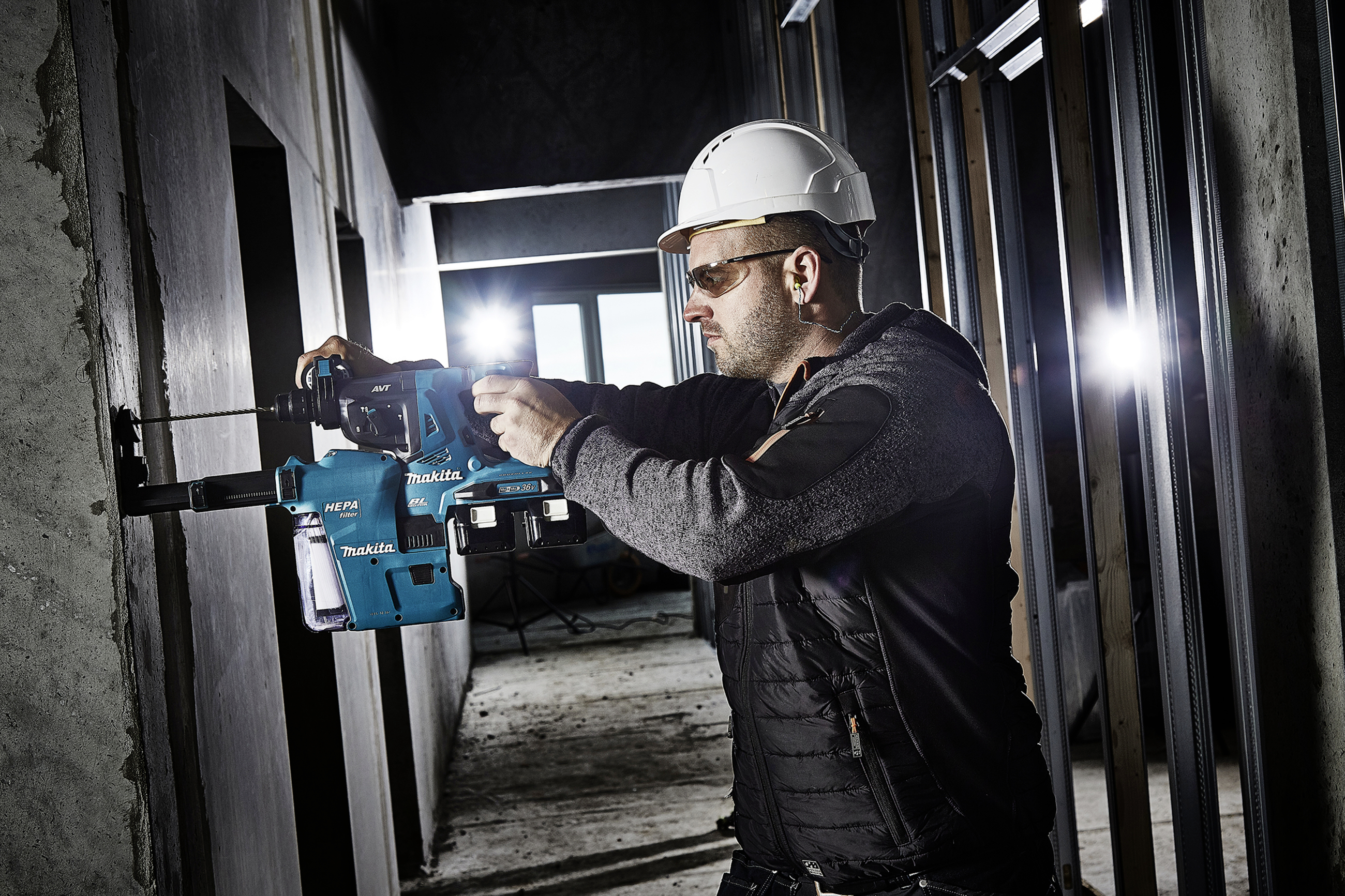 A worker wearing a hard hat and safety glasses is drilling into a concrete wall with an electric power tool in an unfinished building.
