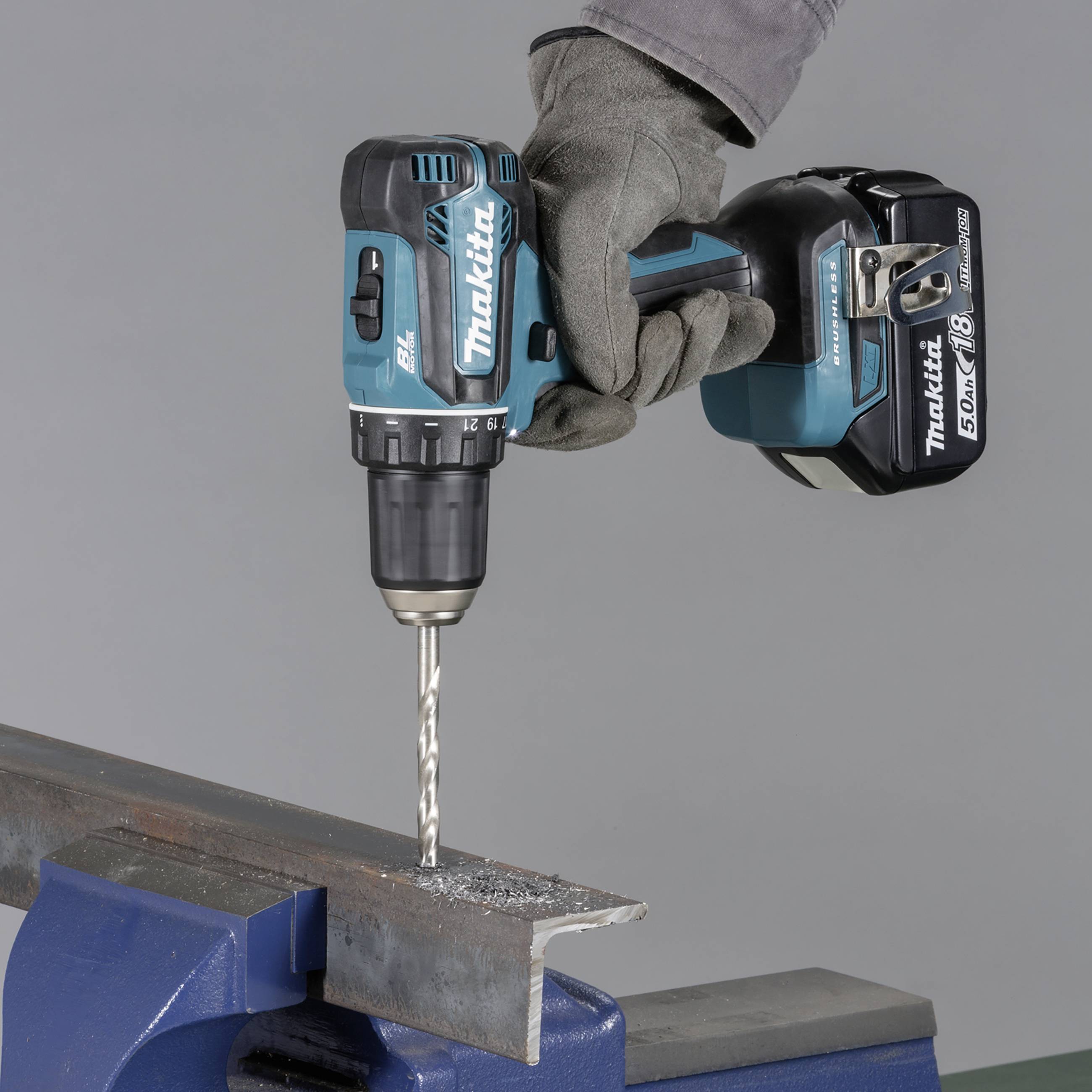 A hand is holding a cordless drill, drilling into metal secured in a vice.