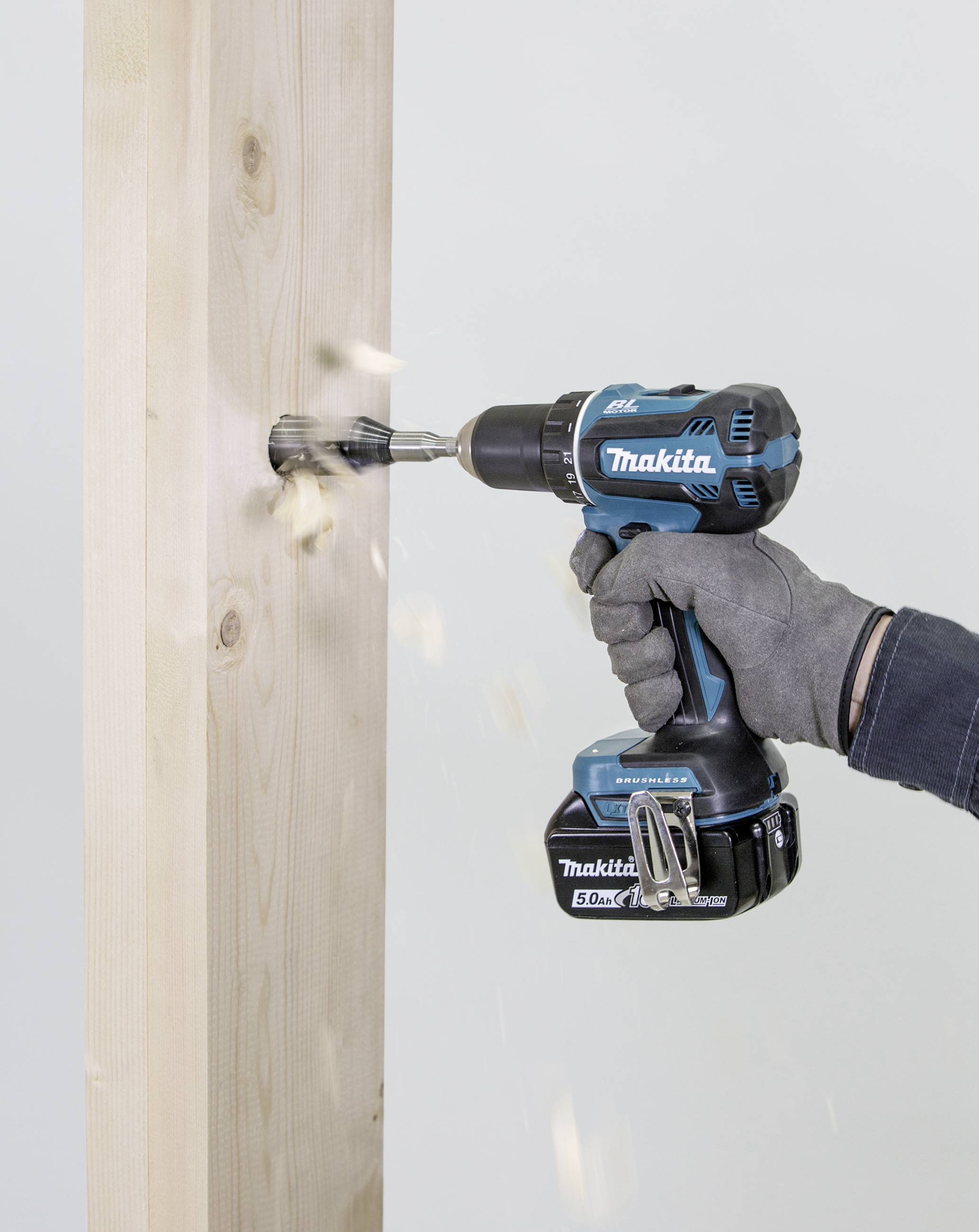 A person is drilling a hole into a vertical wooden post using a blue cordless drill. Wood shavings are flying from the drill hole.