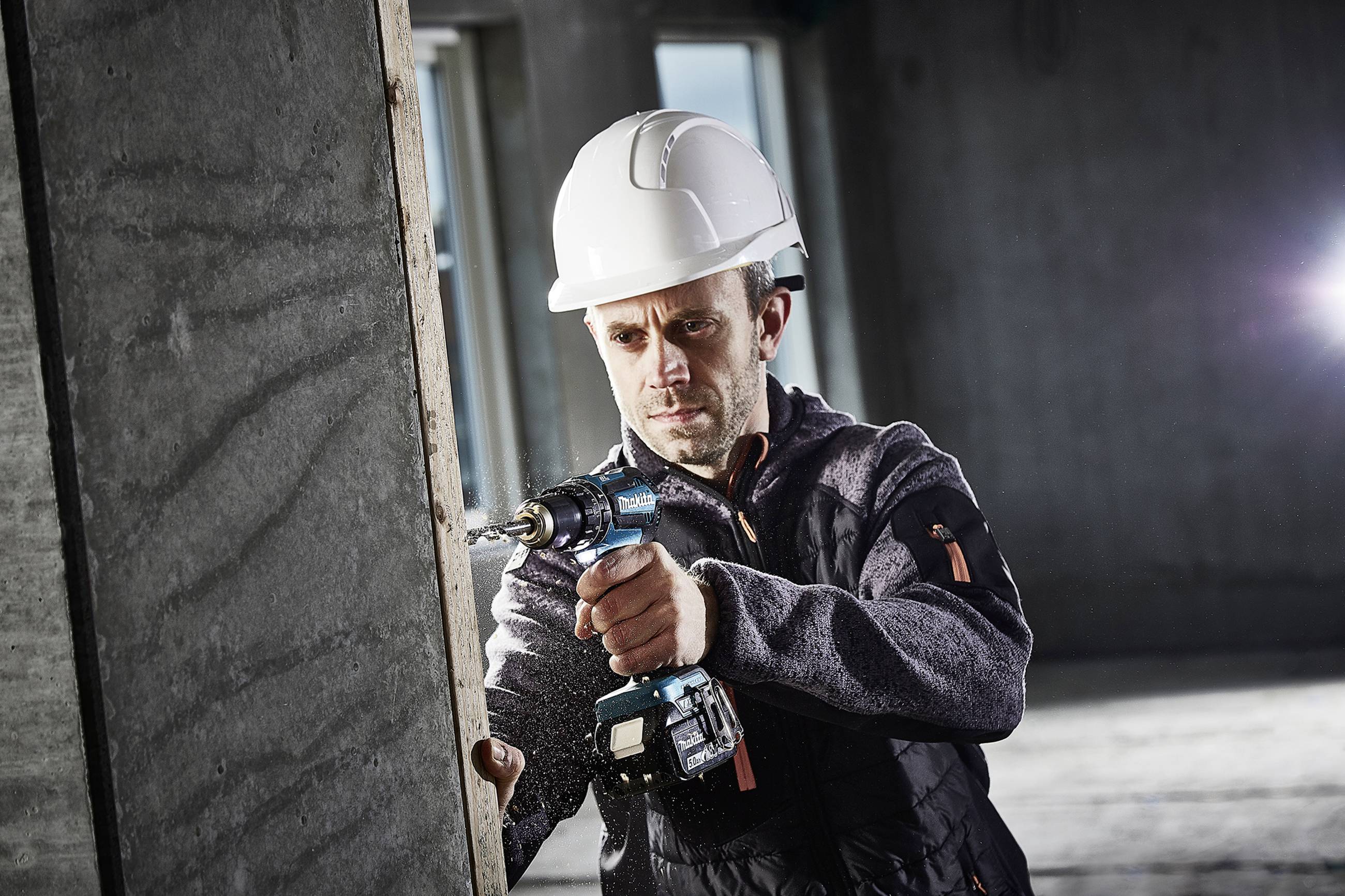 A worker wearing a hard hat is drilling a hole in a concrete wall on a construction site using an electric drill.