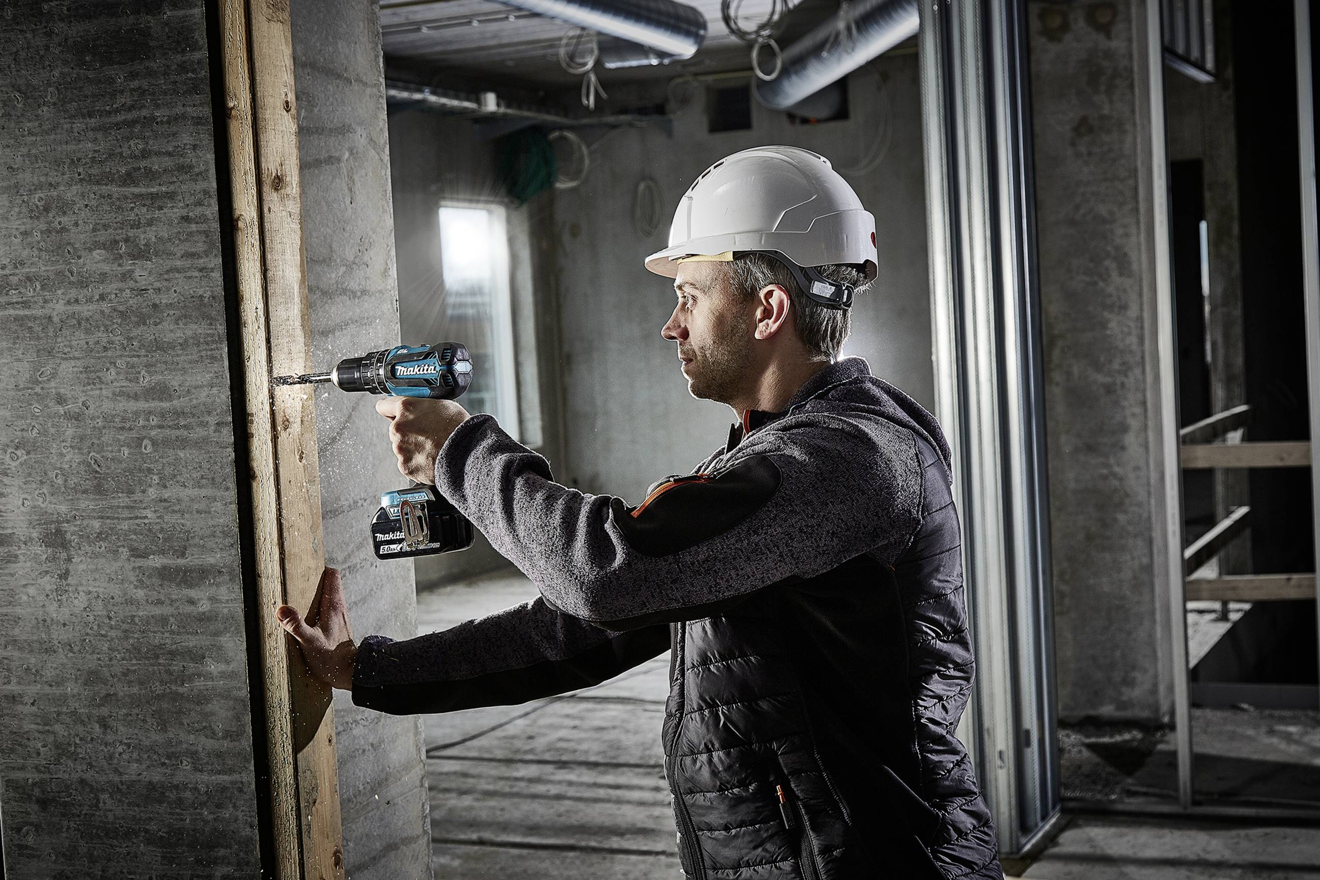 A construction worker wearing a hard hat and workwear is using a cordless drill to bore a screw into a wooden wall in an unfinished building.