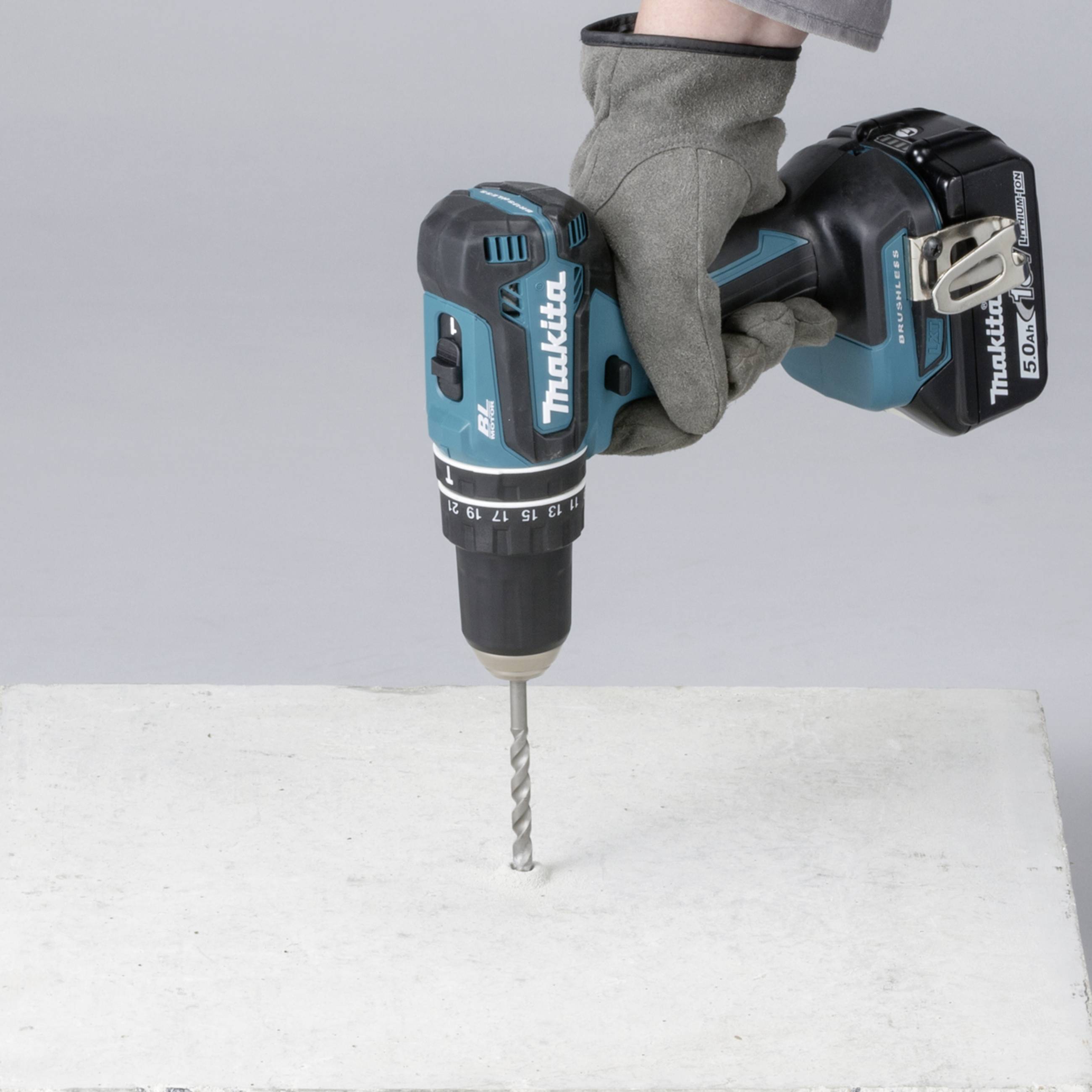 A hand wearing a grey glove is holding a blue cordless drill, drilling into a white concrete slab.