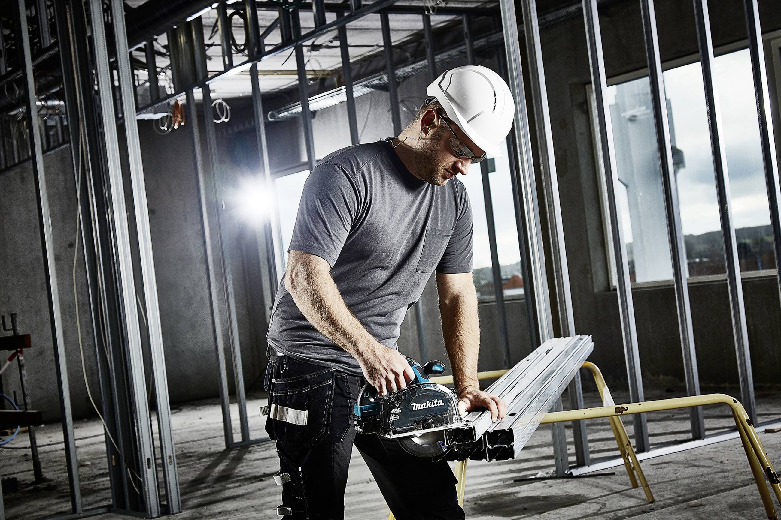 A worker wearing a hard hat is using an electric saw to cut metal struts in an unfinished building. Construction site in the background.