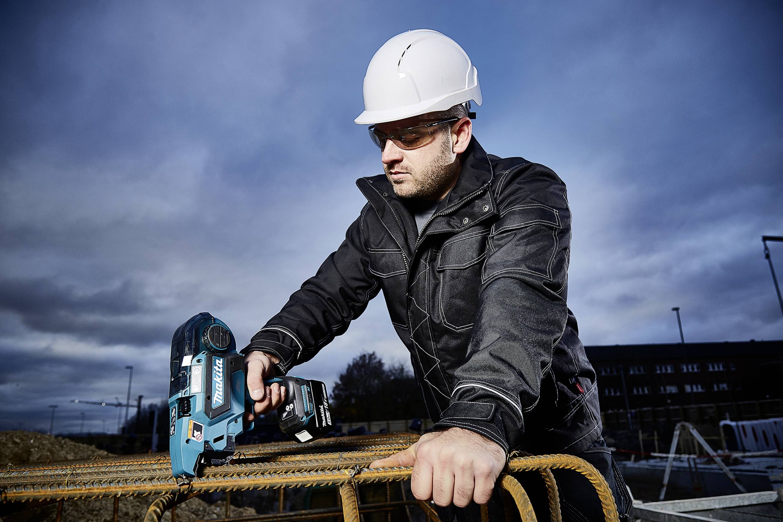 A man wearing a hard hat and safety glasses is working on a construction site, holding an electric tool and leaning over steel reinforcing bars.