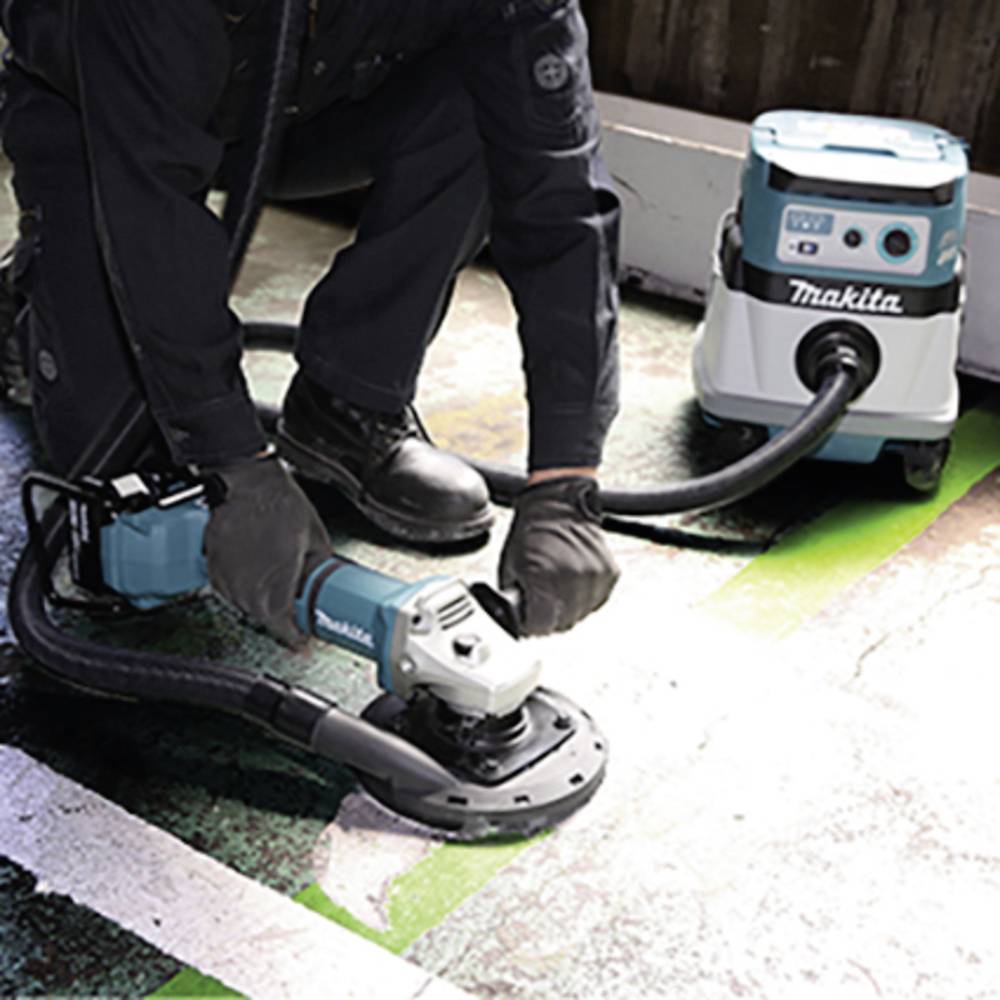 A person is using an electric grinding machine with an attached vacuum cleaner on a floor. In the background, a Makita vacuum cleaner.