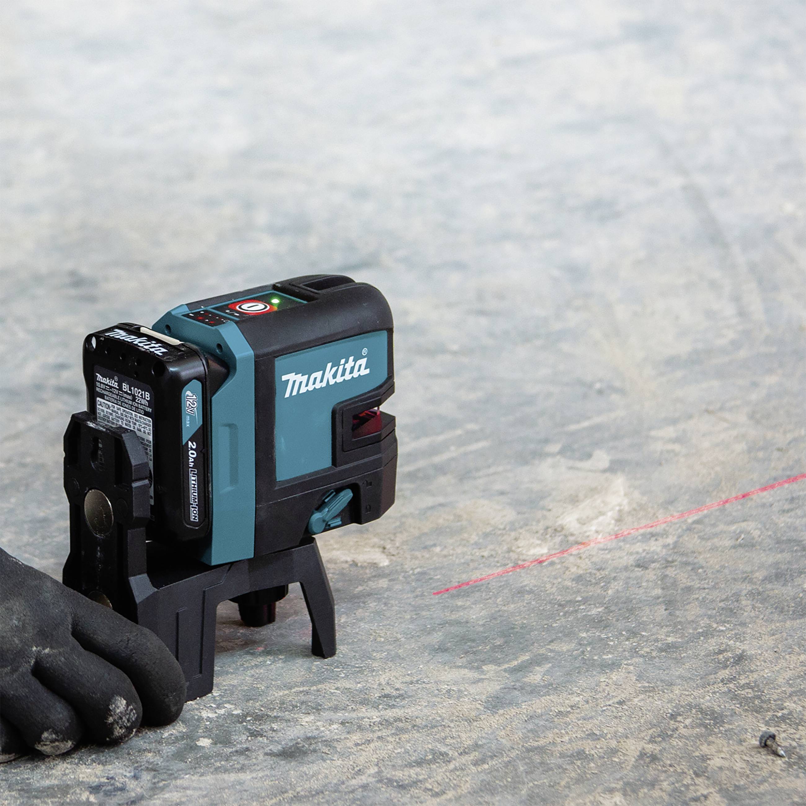 A hand wearing a black glove beside a green and black laser measuring device on a concrete floor. The device is projecting a red laser line.