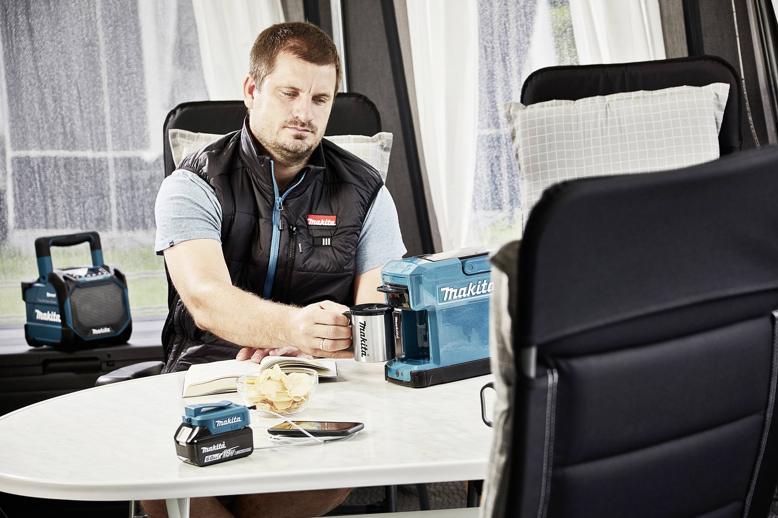 A man is sitting at a table indoors, making coffee with a Makita coffee machine. Snacks and a mobile phone are lying on the table.
