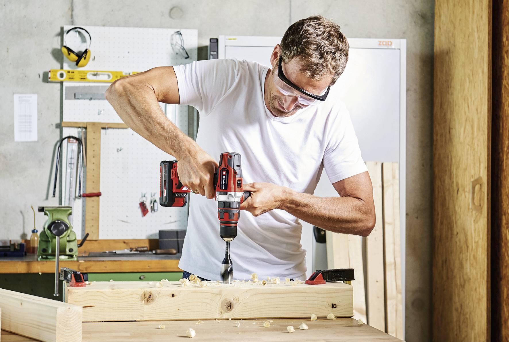 A man is drilling into a wooden board with an electric drill in a workshop. He is wearing safety glasses and working with concentration.