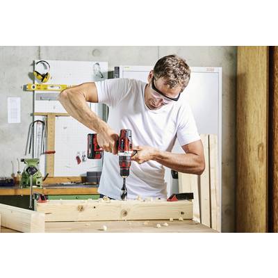 A man is drilling into a wooden board with an electric drill in a workshop. He is wearing safety glasses and working with concentration.
